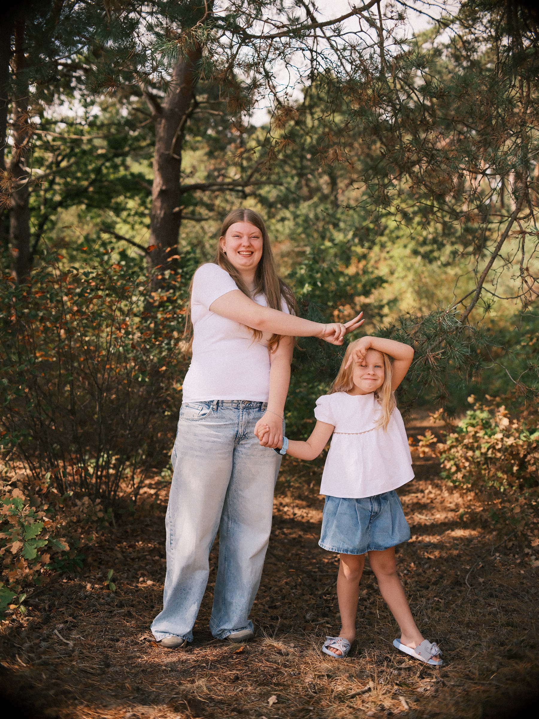 Teenage girl pointing at her little sister's head in a playful height game on a path of pine needles, both in white and denim