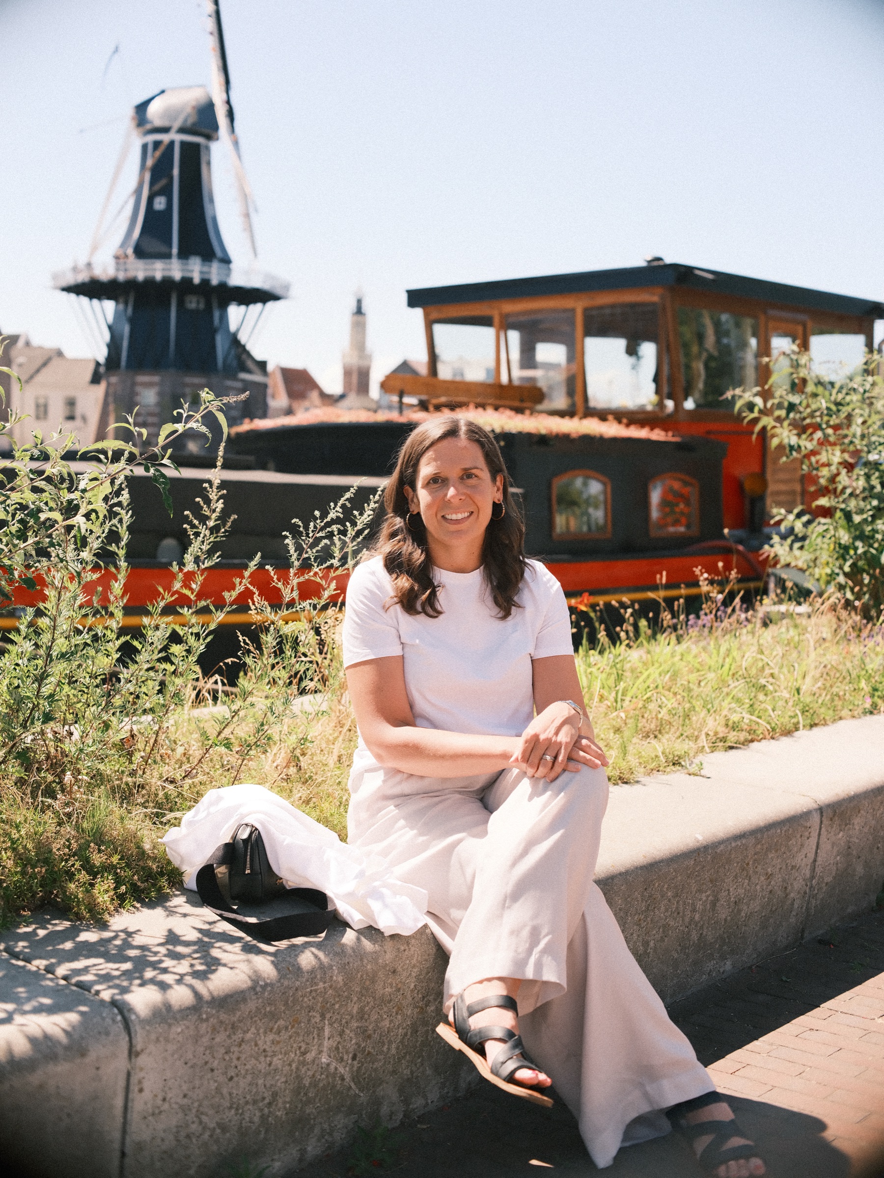Vrouw in wit T-shirt zit op een lage betonnen rand in het gras, haar blazer naast zich en De Adriaan achter het schip