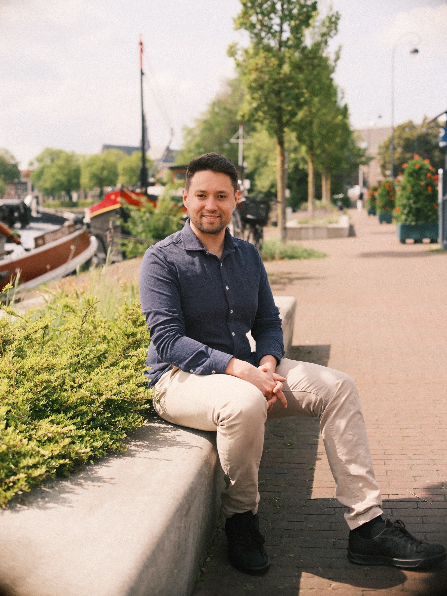 Lachende man in donkerblauw overhemd zit op een laag muurtje bij afgemeerde boten, met een klinkerpad en jonge bomen erachter