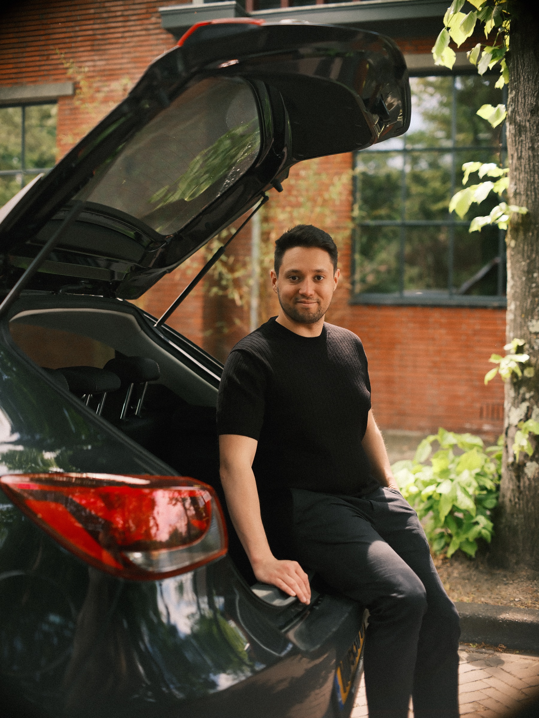 Man zit op de rand van de open achterklep van zijn auto, met het rode achterlicht vooraan en een bakstenen gebouw met groen erachter