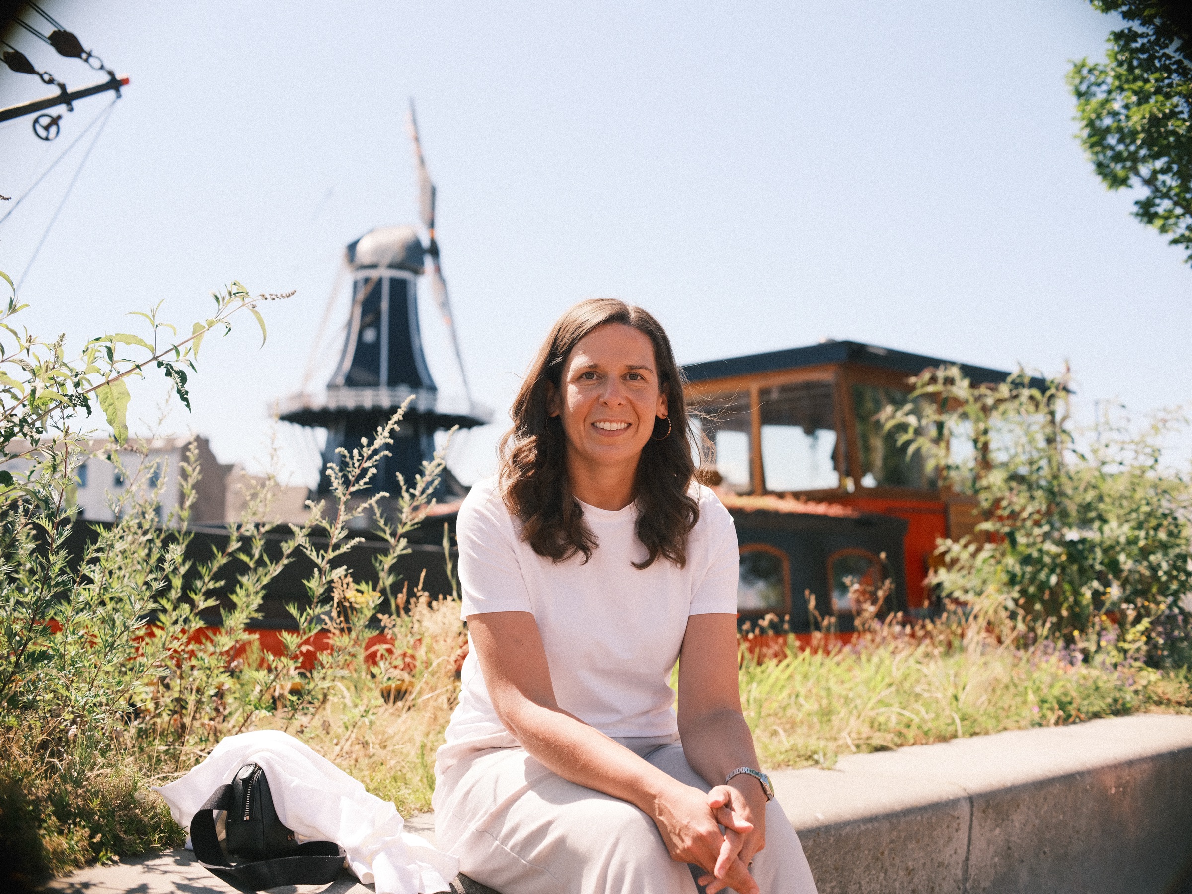 Lachende vrouw in het wit op een lage rand tussen wilde grassen, met molen De Adriaan en de houten stuurhut van het schip