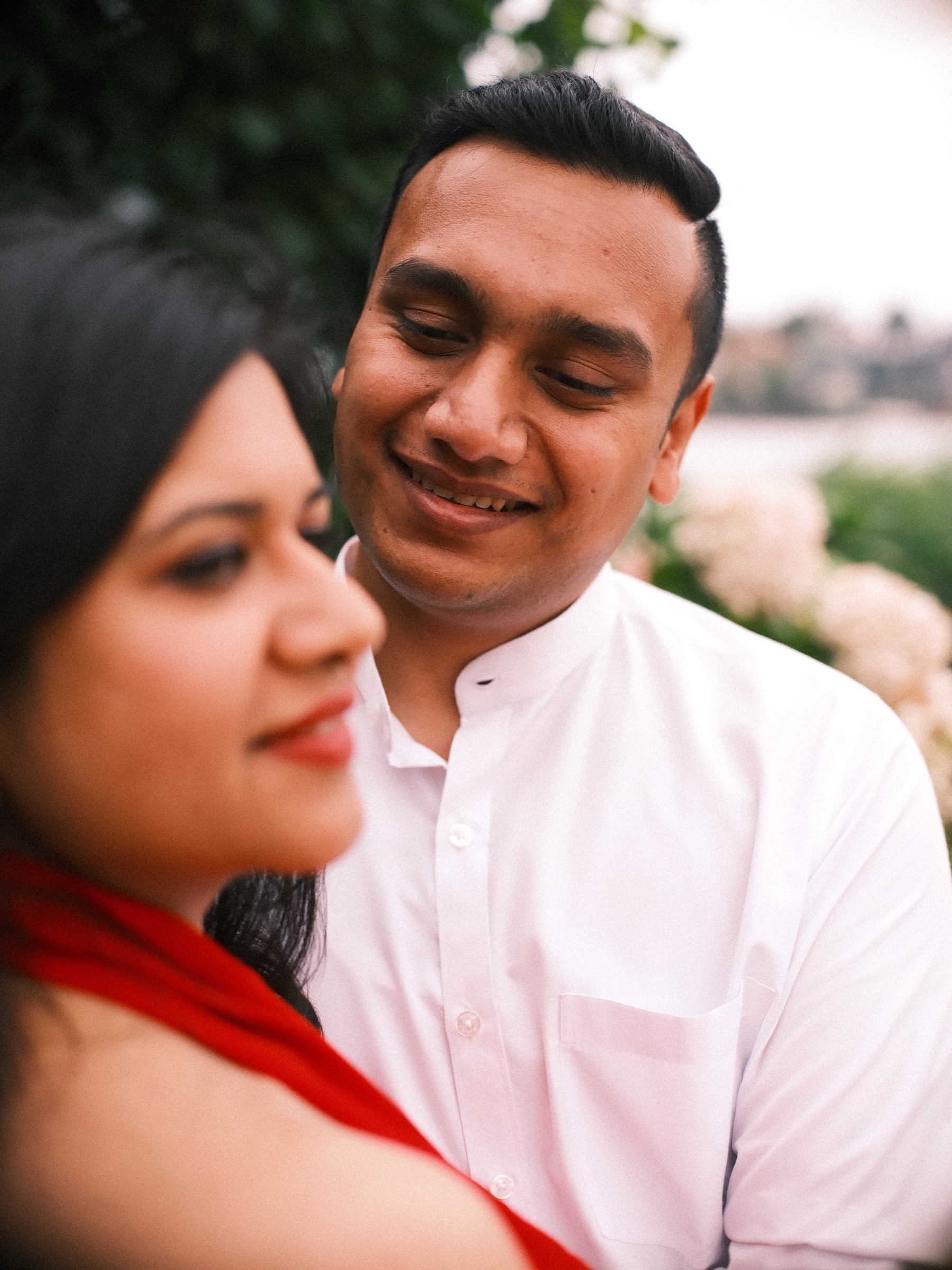 Close frame with her face soft in the foreground and his smile sharp behind it, a white shirt and a hint of crimson against blurred flowers