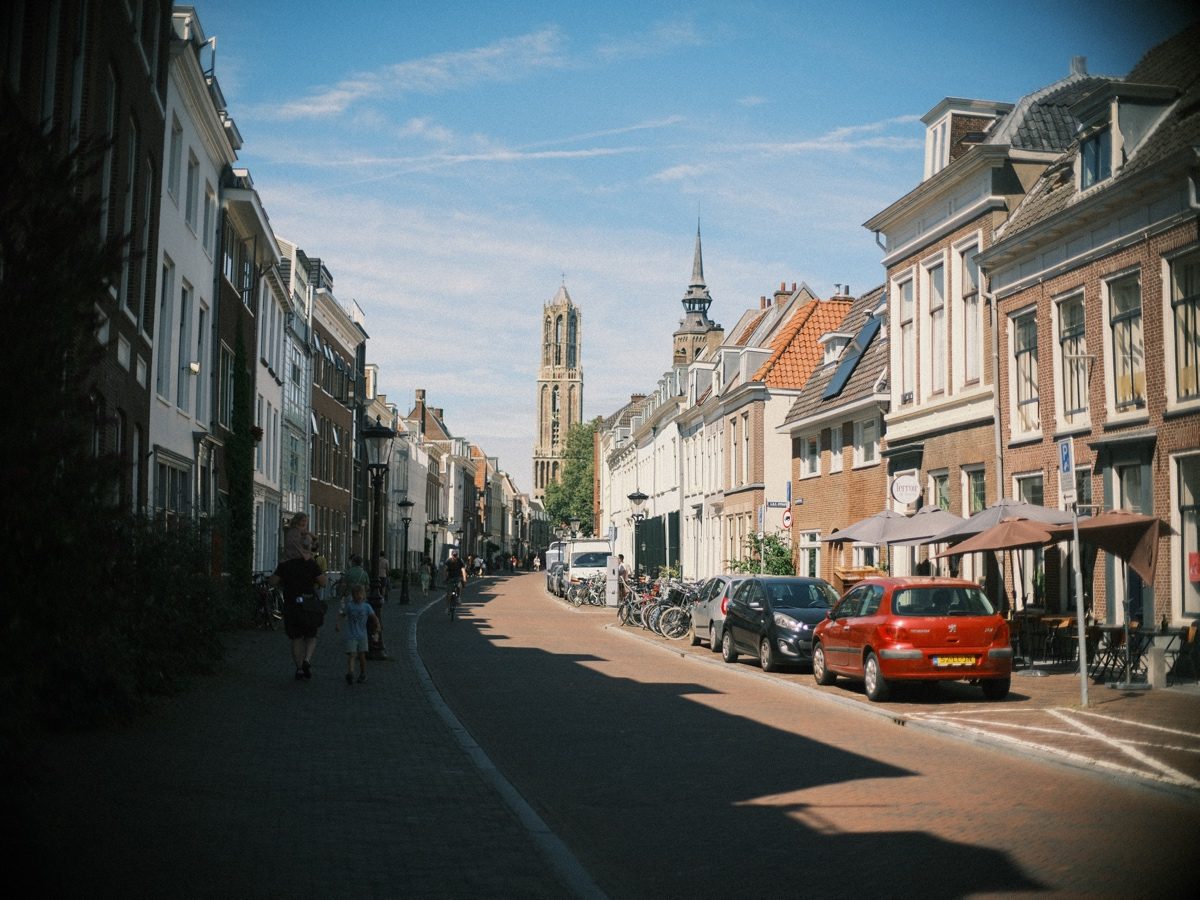 Sunny Utrecht street with old house fronts and parked cars, the Dom Tower and a church spire rising at its end