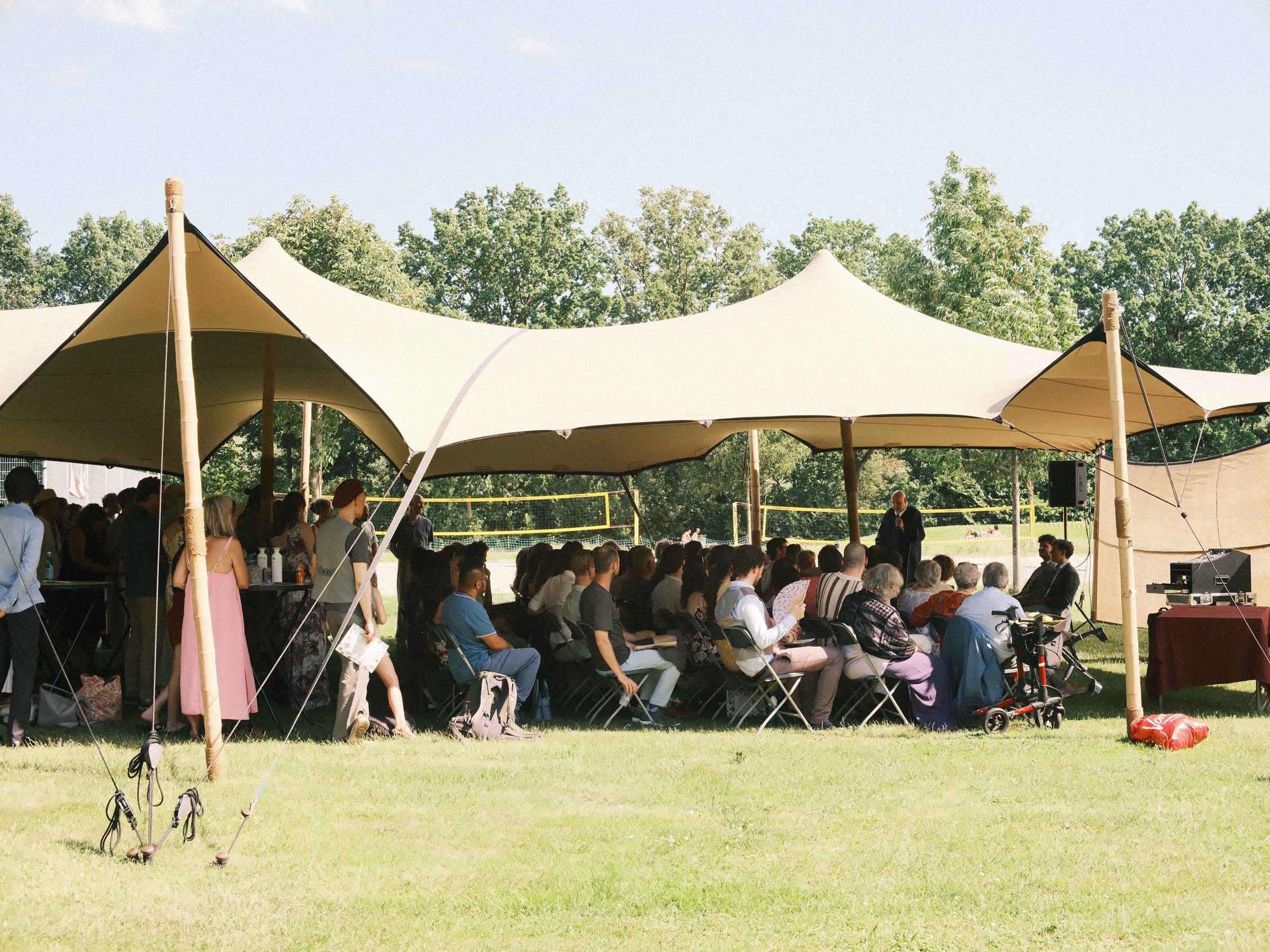 Sand-colored stretch tents on a sunny lawn in Utrecht, wedding guests seated in the shade and tall trees behind them