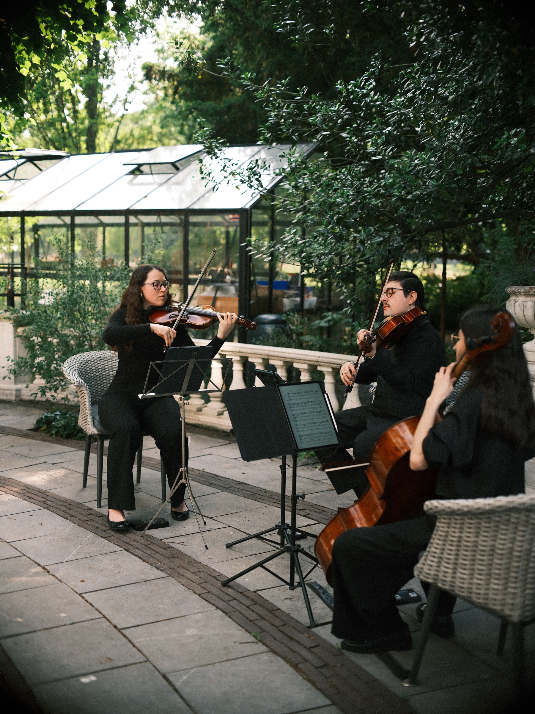 Strijktrio met een cello speelt op een stenen terras bij een witte balustrade, met een glazen kas tussen de bomen erachter