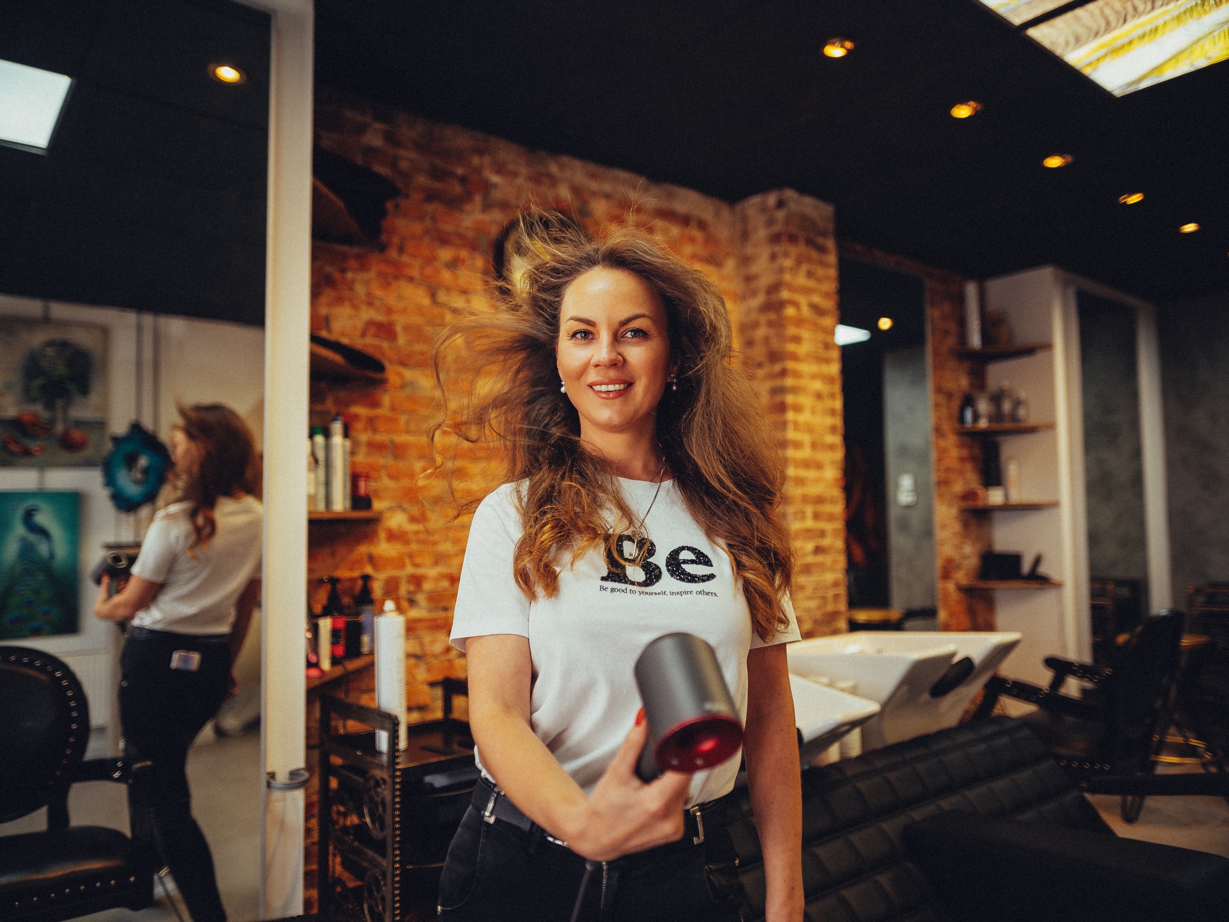 Eigenaar in een wit Be-shirt met een föhn in haar beautystudio, haar lange haar waait op voor een rode bakstenen muur