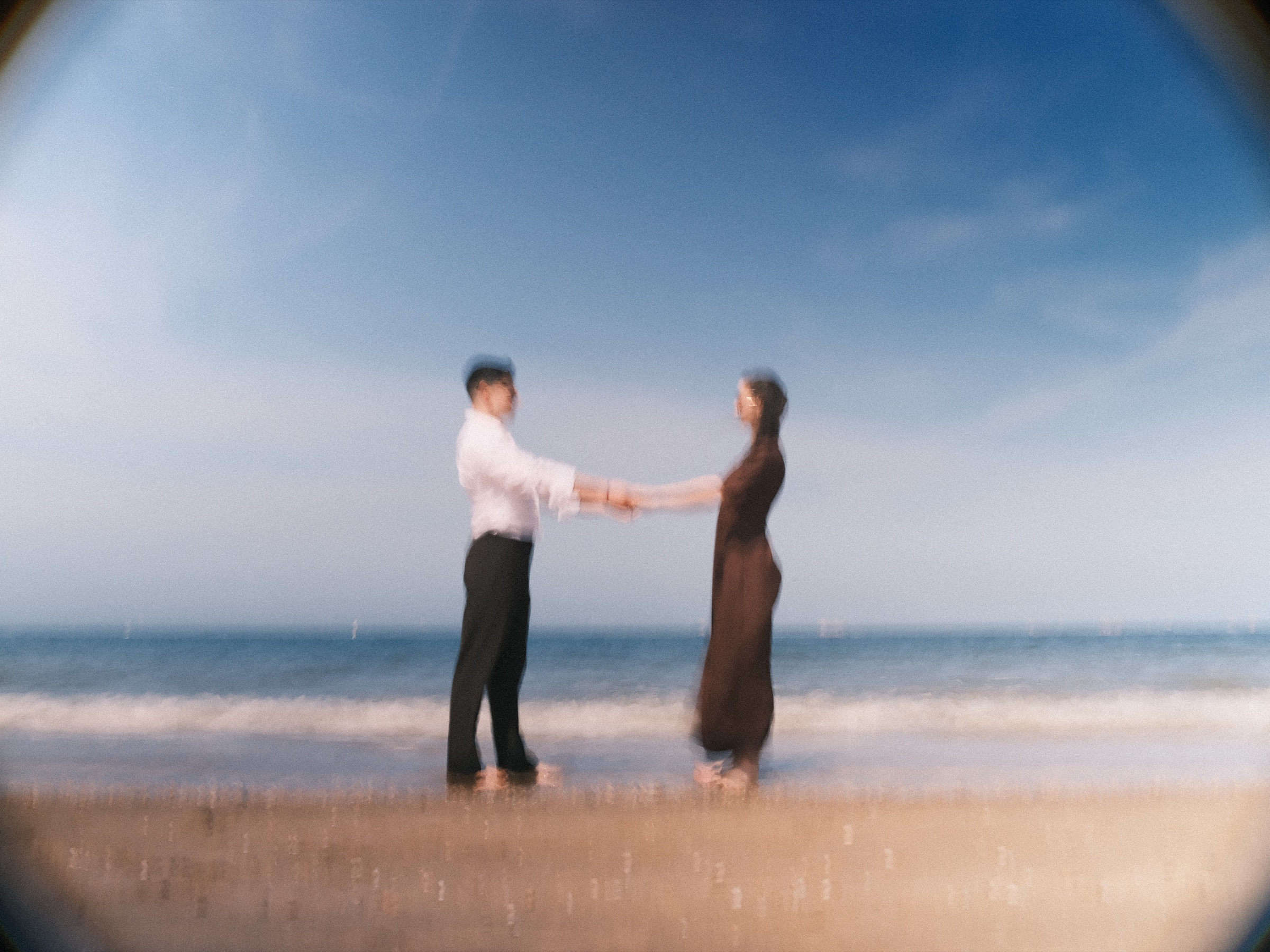 Soft swirling blur of the couple holding hands at the water's edge, the sea and the sky melting together around them