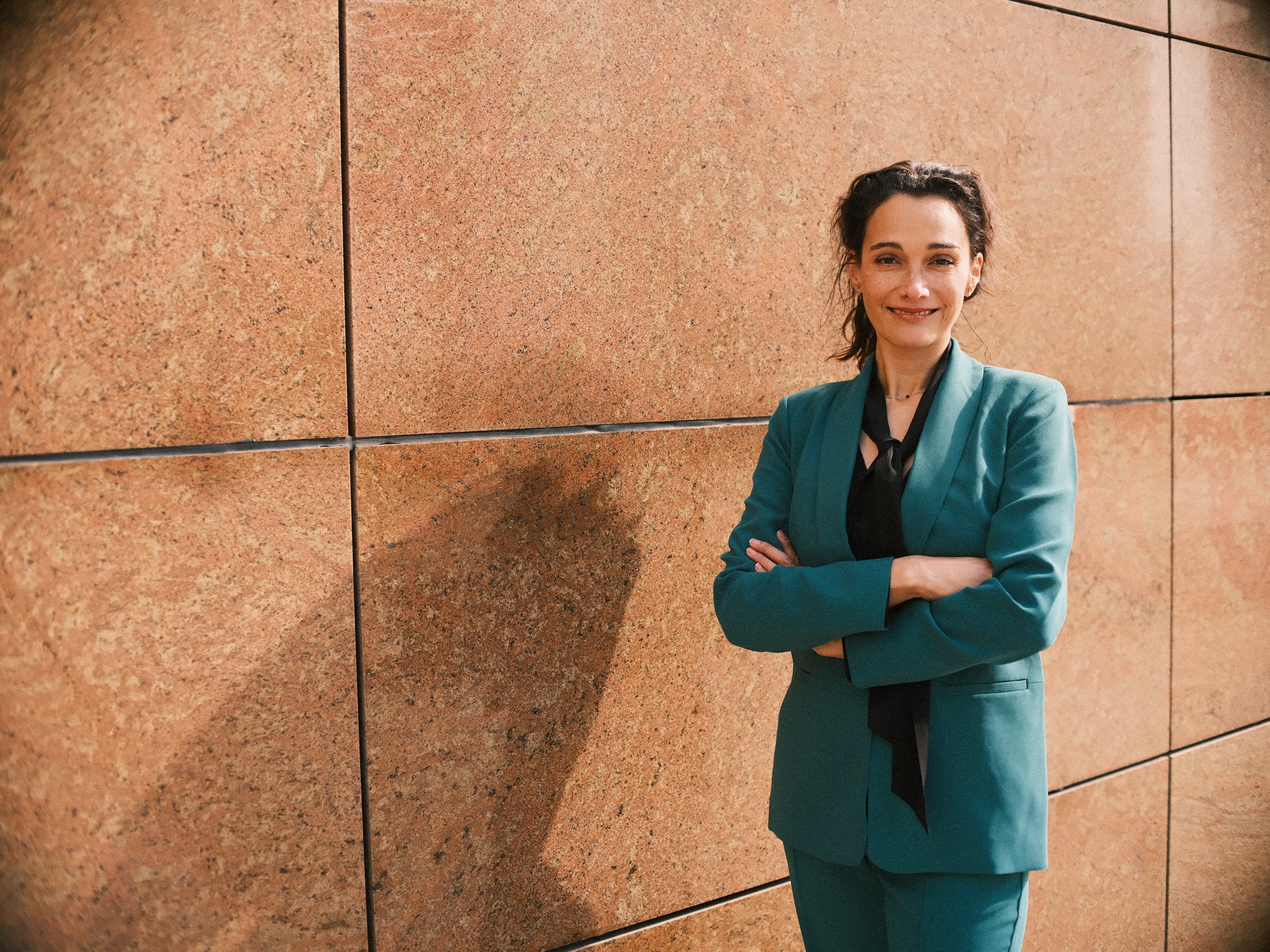 Vrouw in een petrolblauw pak met de armen over elkaar lacht tegen een warme stenen muur in de felle maartzon van Den Haag