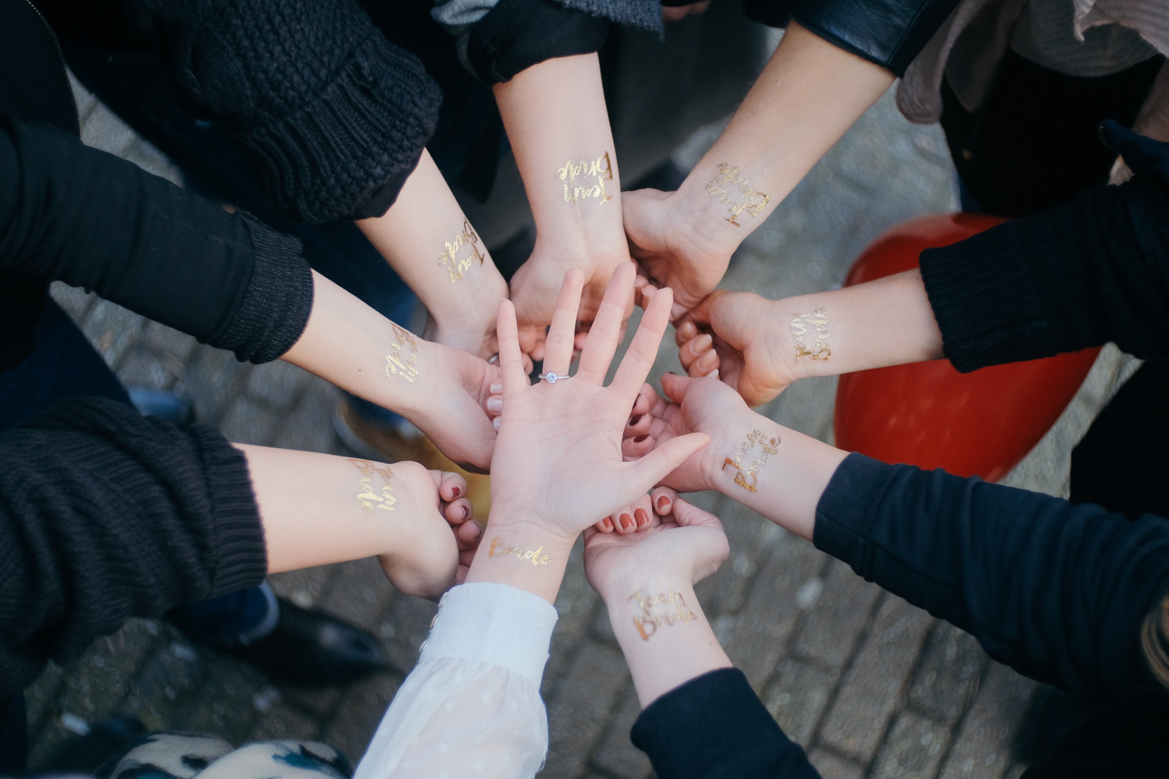 Circle of hands with gold Team Bride tattoos around the bride's open palm and diamond ring, seen from above at the hen party