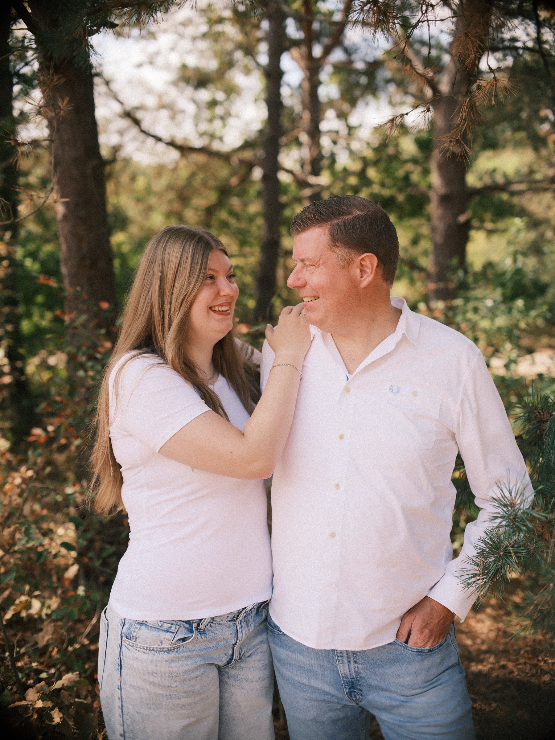 Teenage girl resting a hand on her father's shoulder as they laugh at each other among the pines, both in white tops