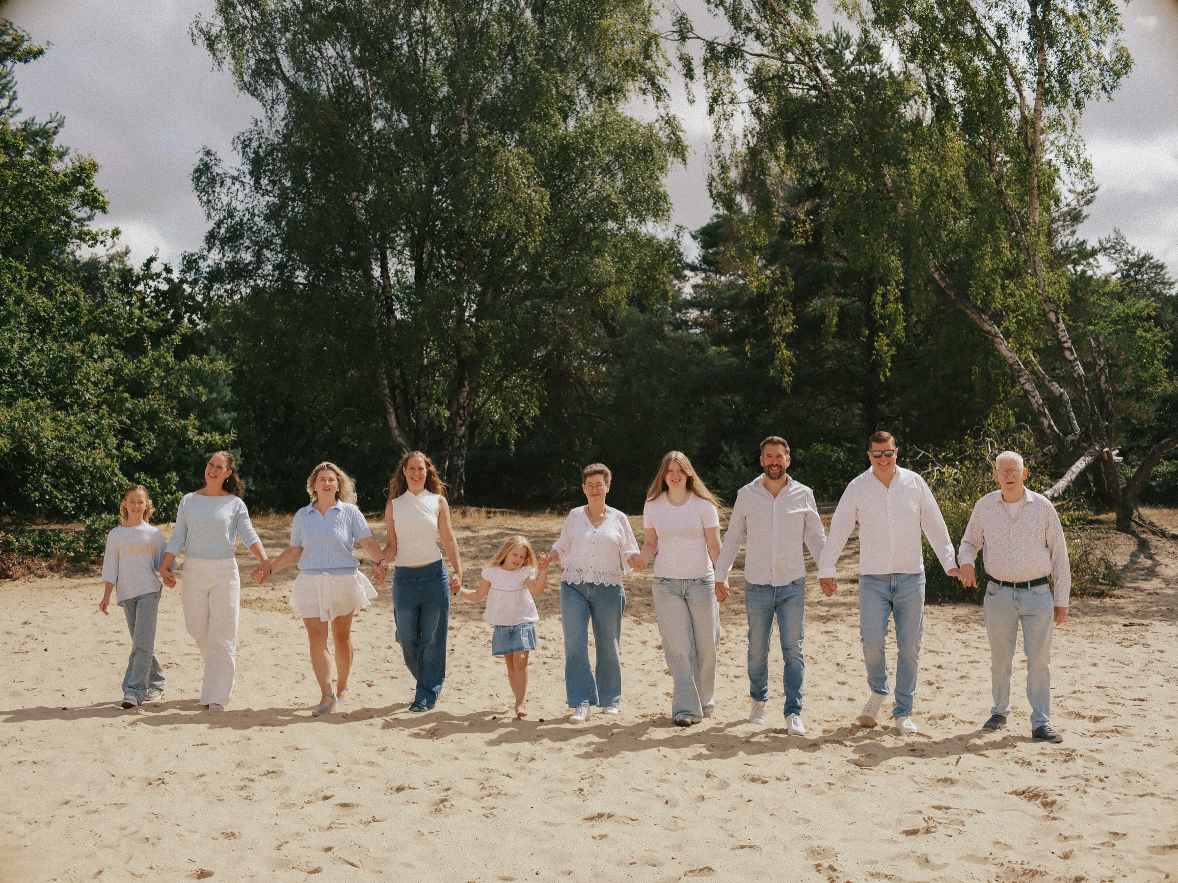 Three generations walking hand in hand across the sand, ten family members in one long line in the dunes with birches behind