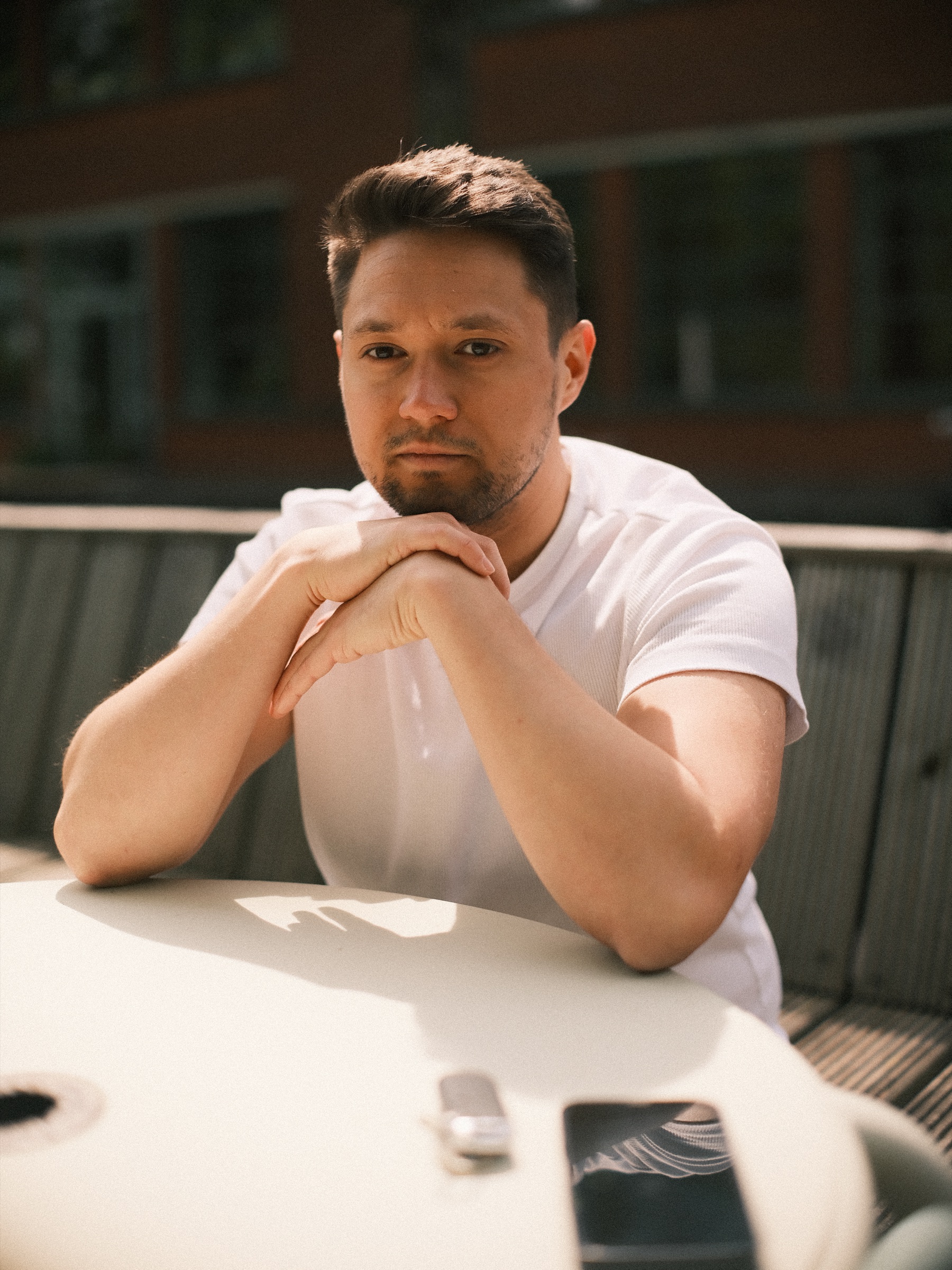 Man in wit T-shirt leunt op een rond wit tafeltje op een zonnig terras, kin op zijn handen en zijn telefoon voor zich