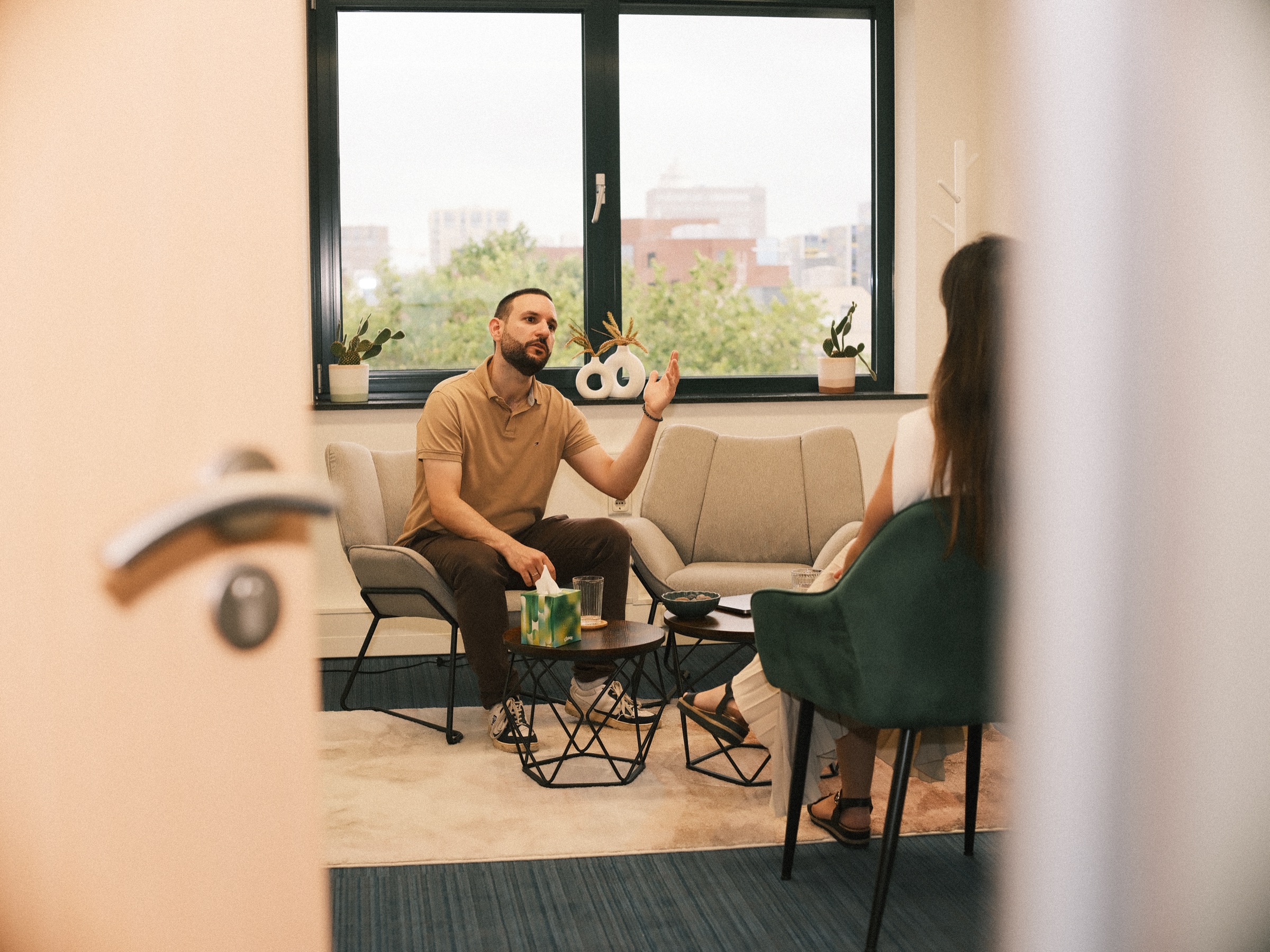Session in a therapy practice seen through a half-open door, a man in a tan polo talking to a woman in a green velvet chair