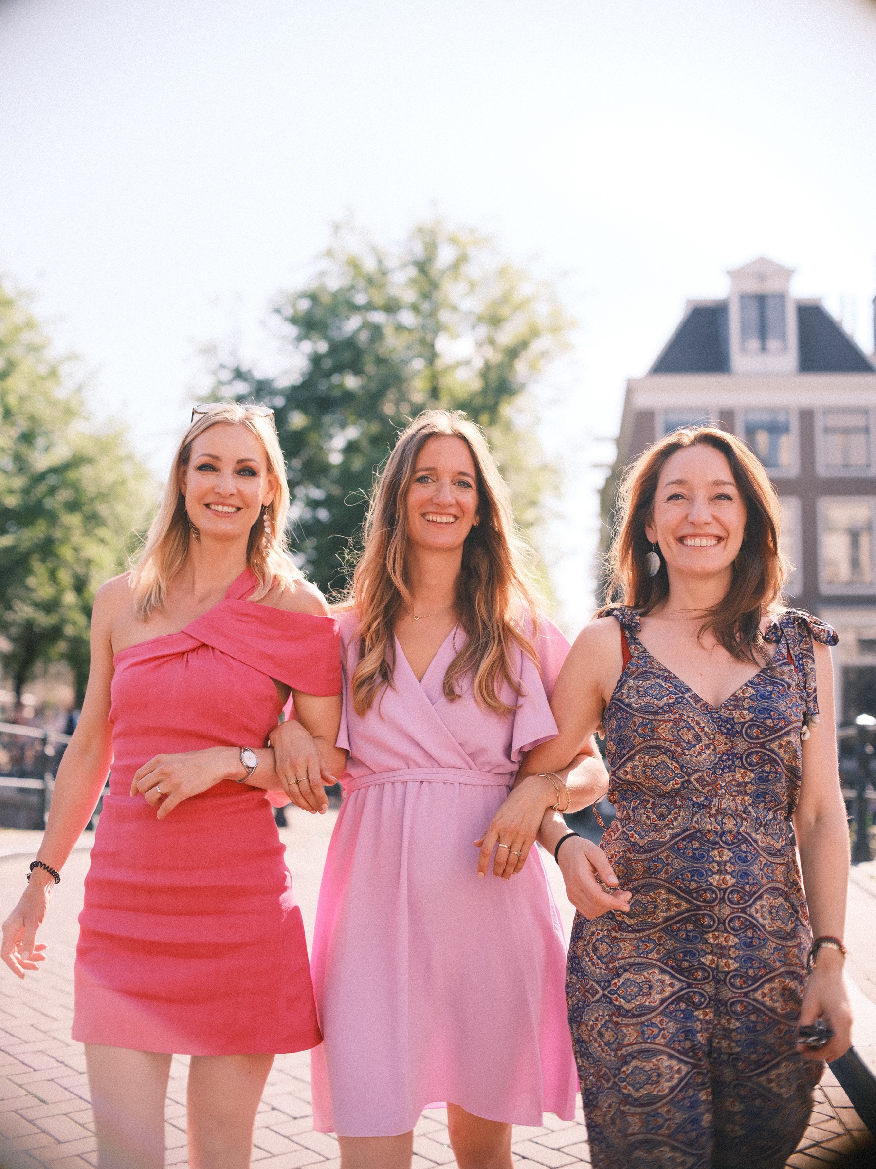 Three women arm in arm on a sunny canal bridge, a bachelorette with twelve friends, the bride-to-be in lilac in the middle