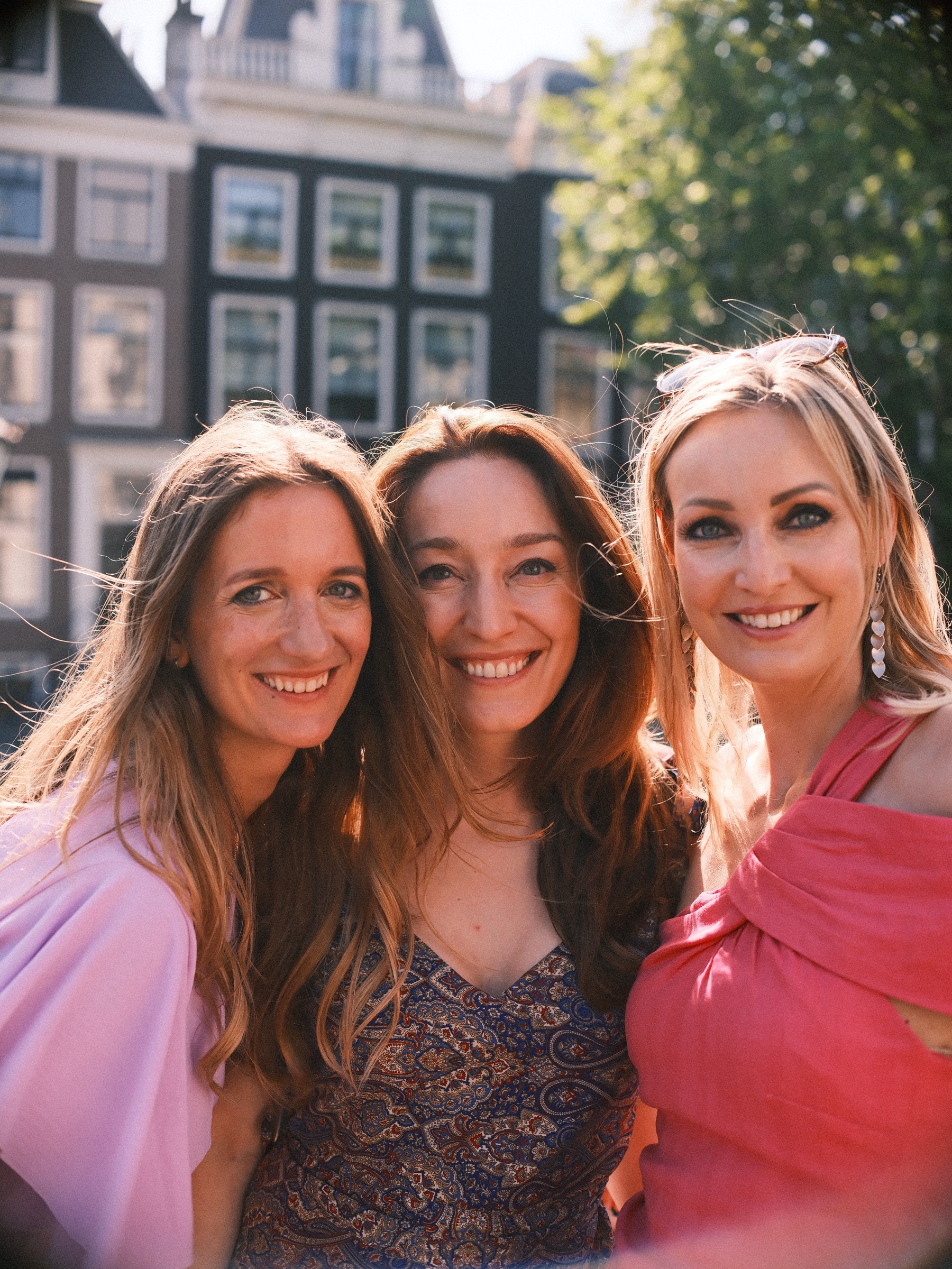 Three women leaning close and smiling, a pink one-shoulder dress on the right and canal house windows behind them