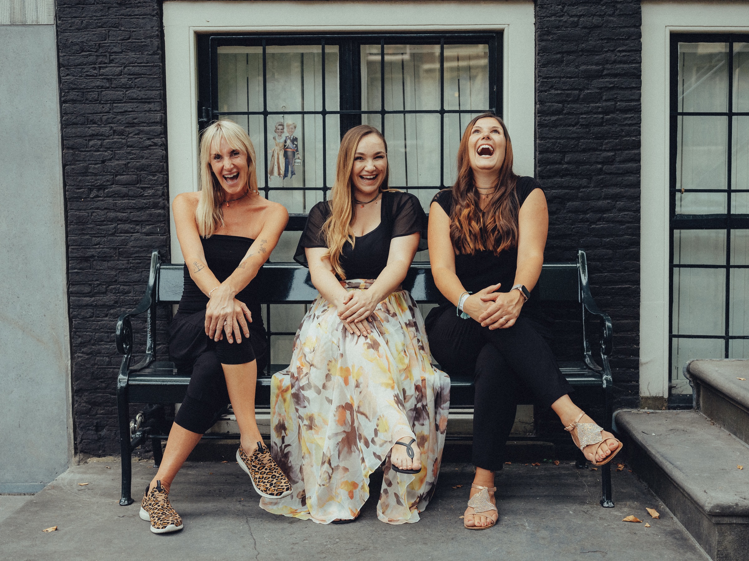 Three friends laughing on a green bench in front of a black brick facade, legs crossed and sandals on
