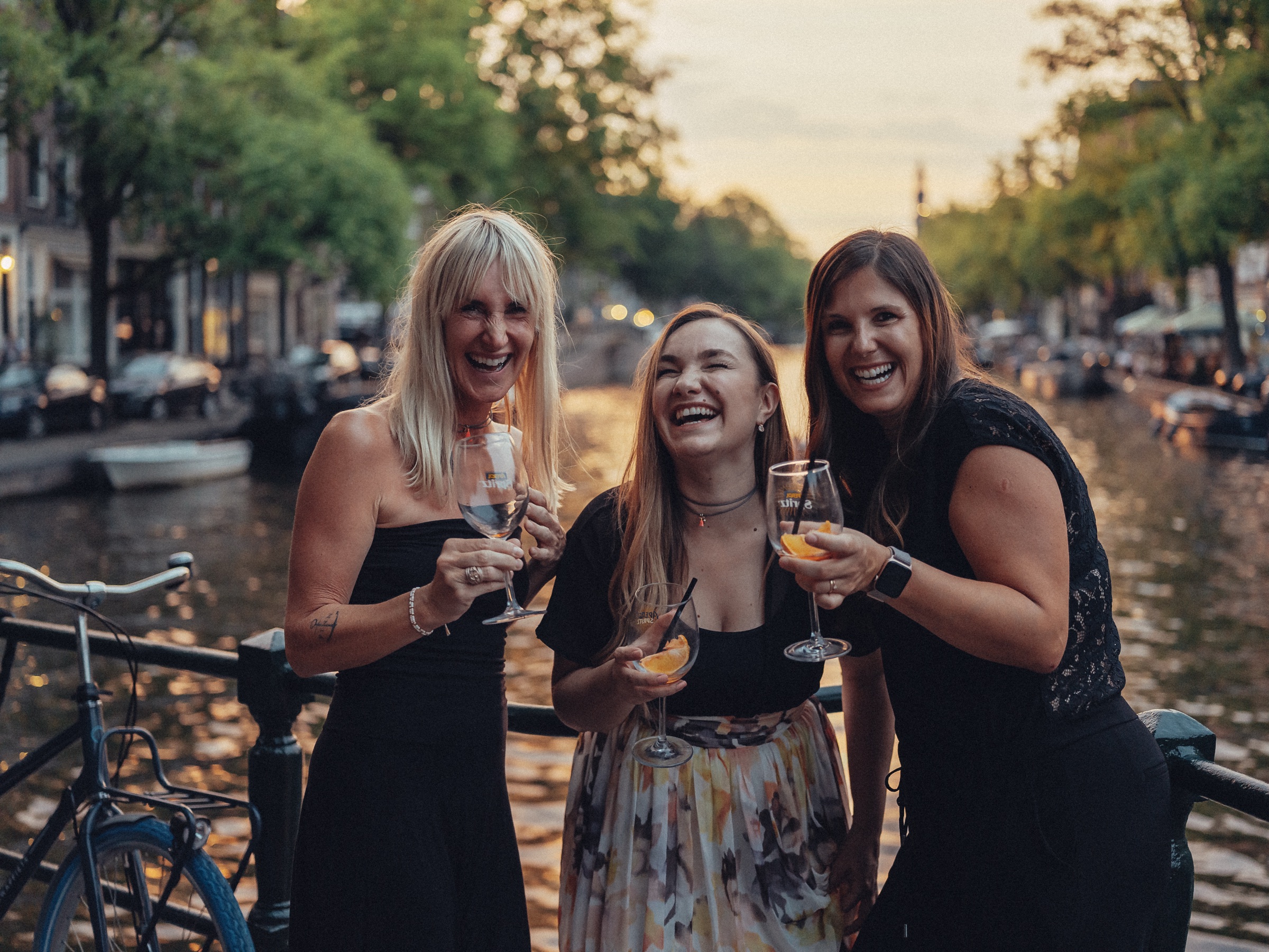 Three friends laughing with their glasses on a canal bridge at sunset, the last light of the bachelorette party on the water
