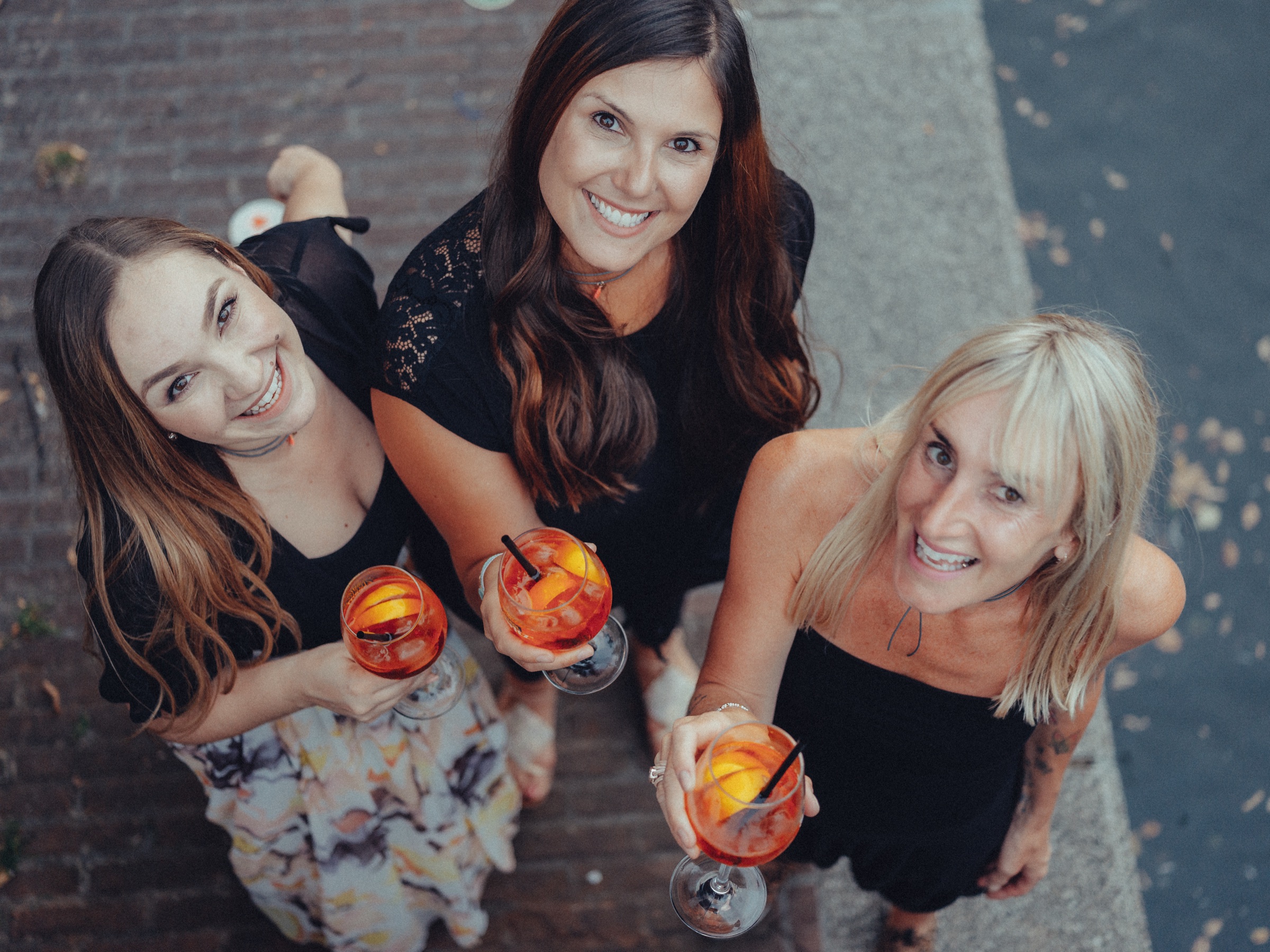 Bachelorette party friends looking up with spritz glasses in hand, seen from above on the brick sidewalk
