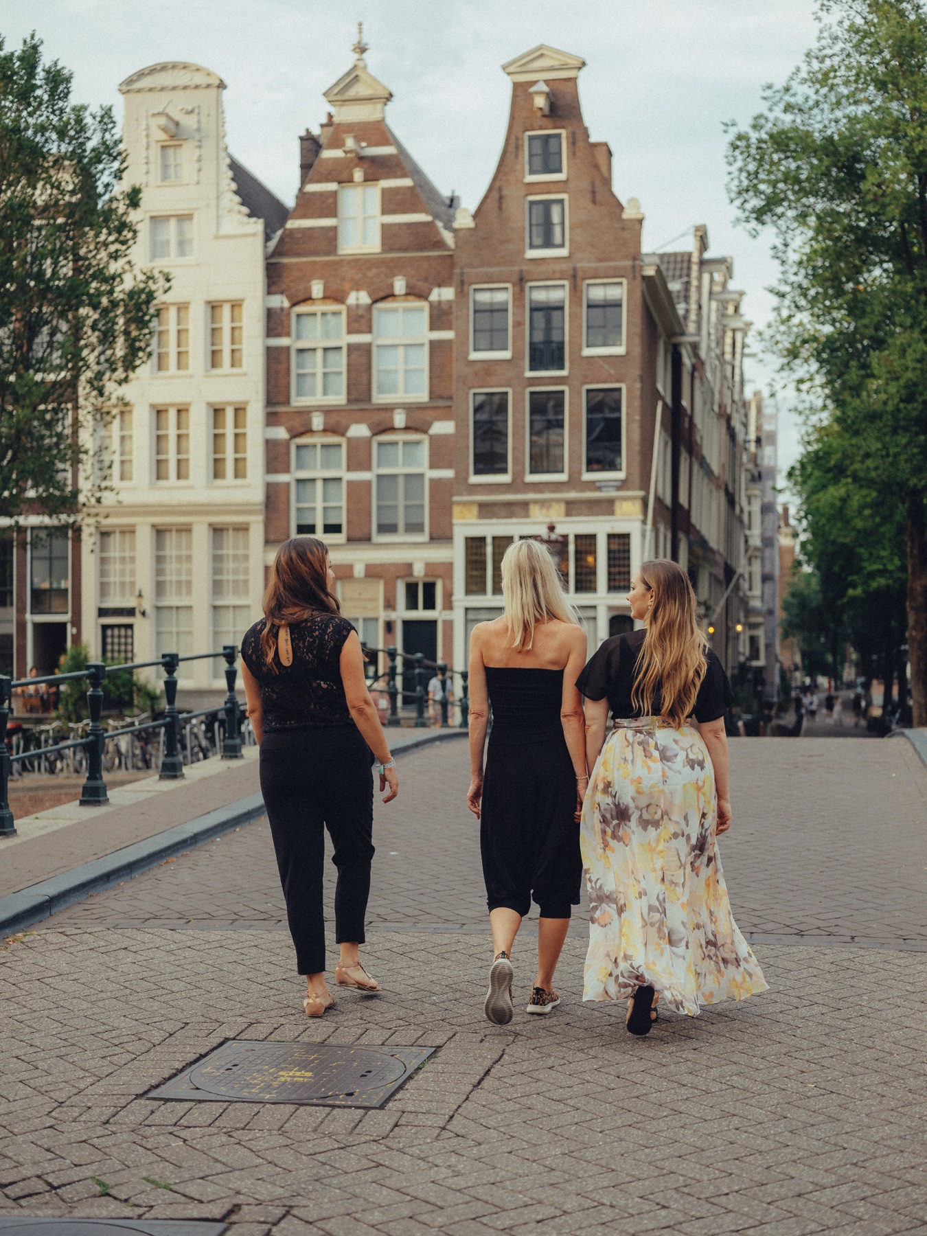 Three friends walking away across a bridge toward tall gabled houses, the floral skirt swinging in the evening light