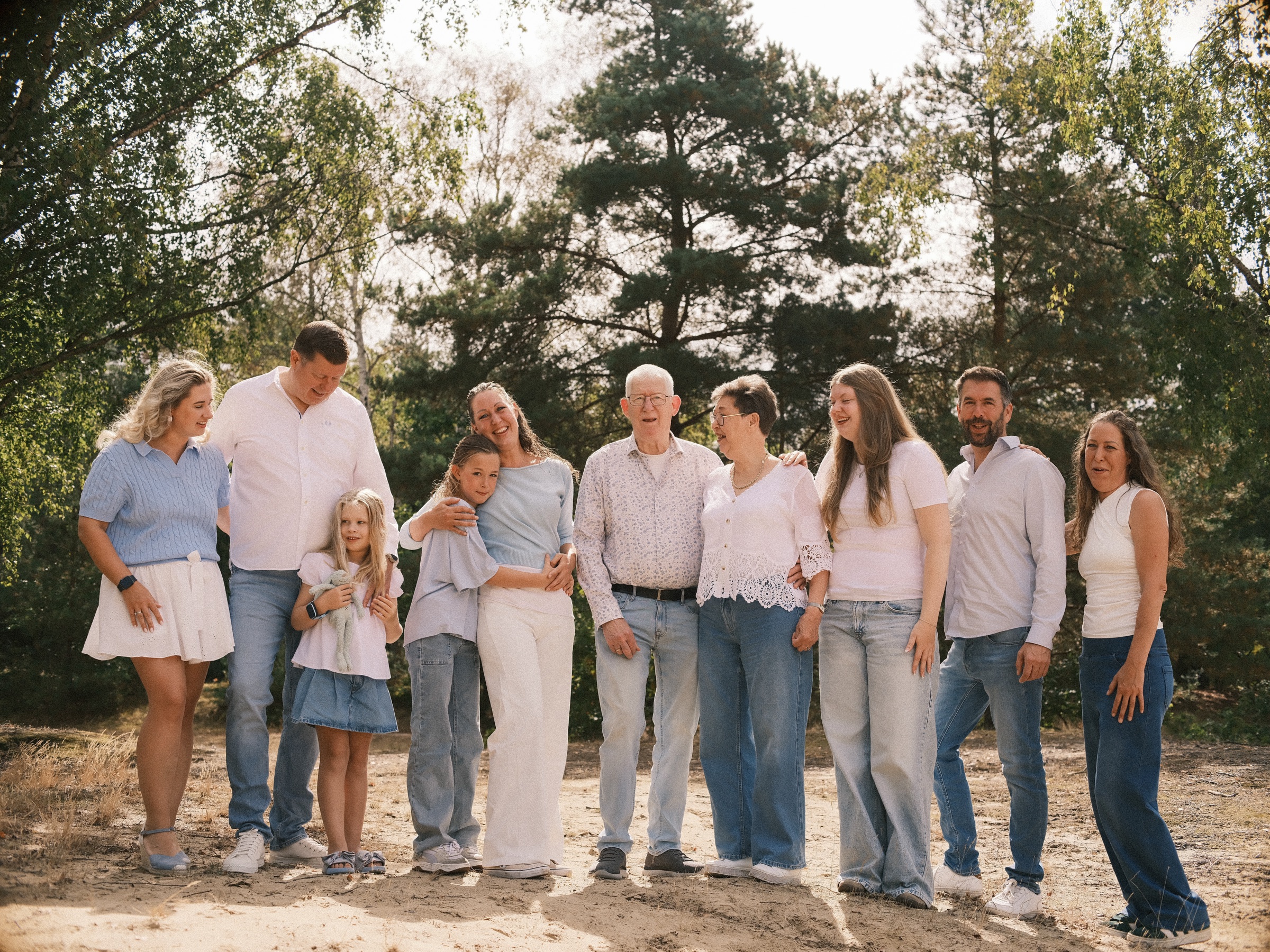 Three generations of one family laughing together on the sand in the dunes, ten people in white, blue and denim before tall pines