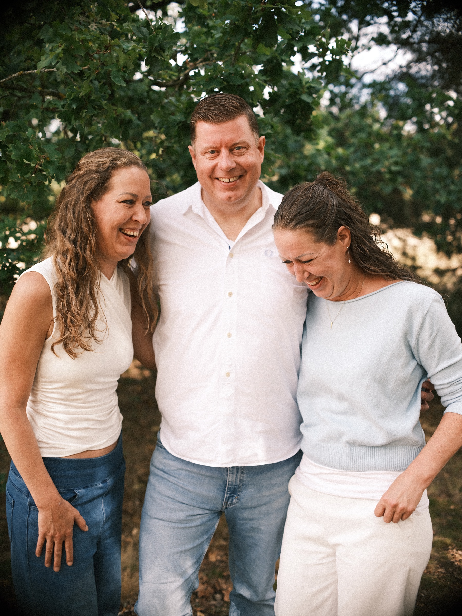 Grown siblings doubled over with laughter in front of green leaves, the brother in a white shirt between his two sisters