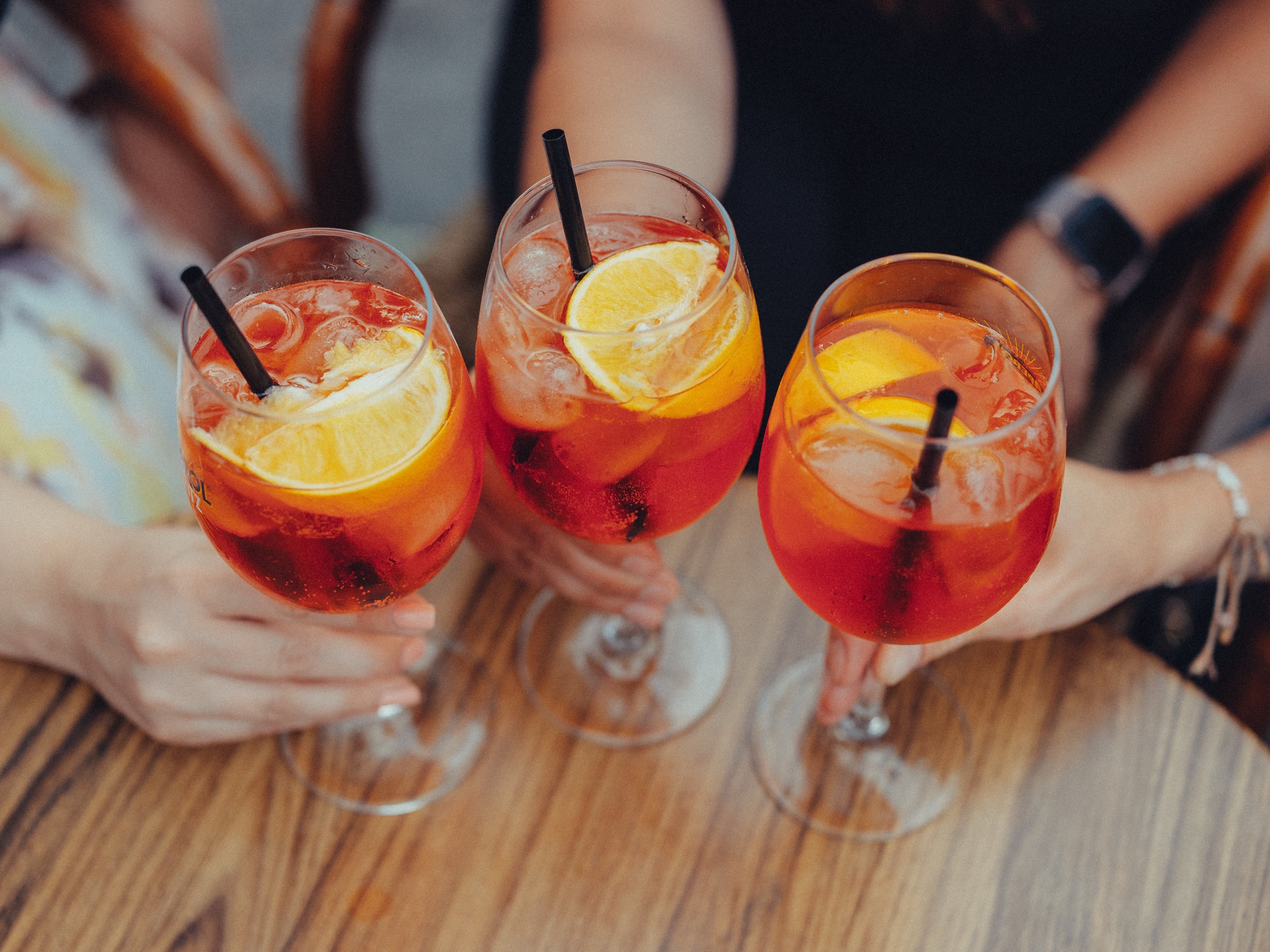 Three glasses of spritz with orange slices and black straws clinking over a wooden café table at the bachelorette party