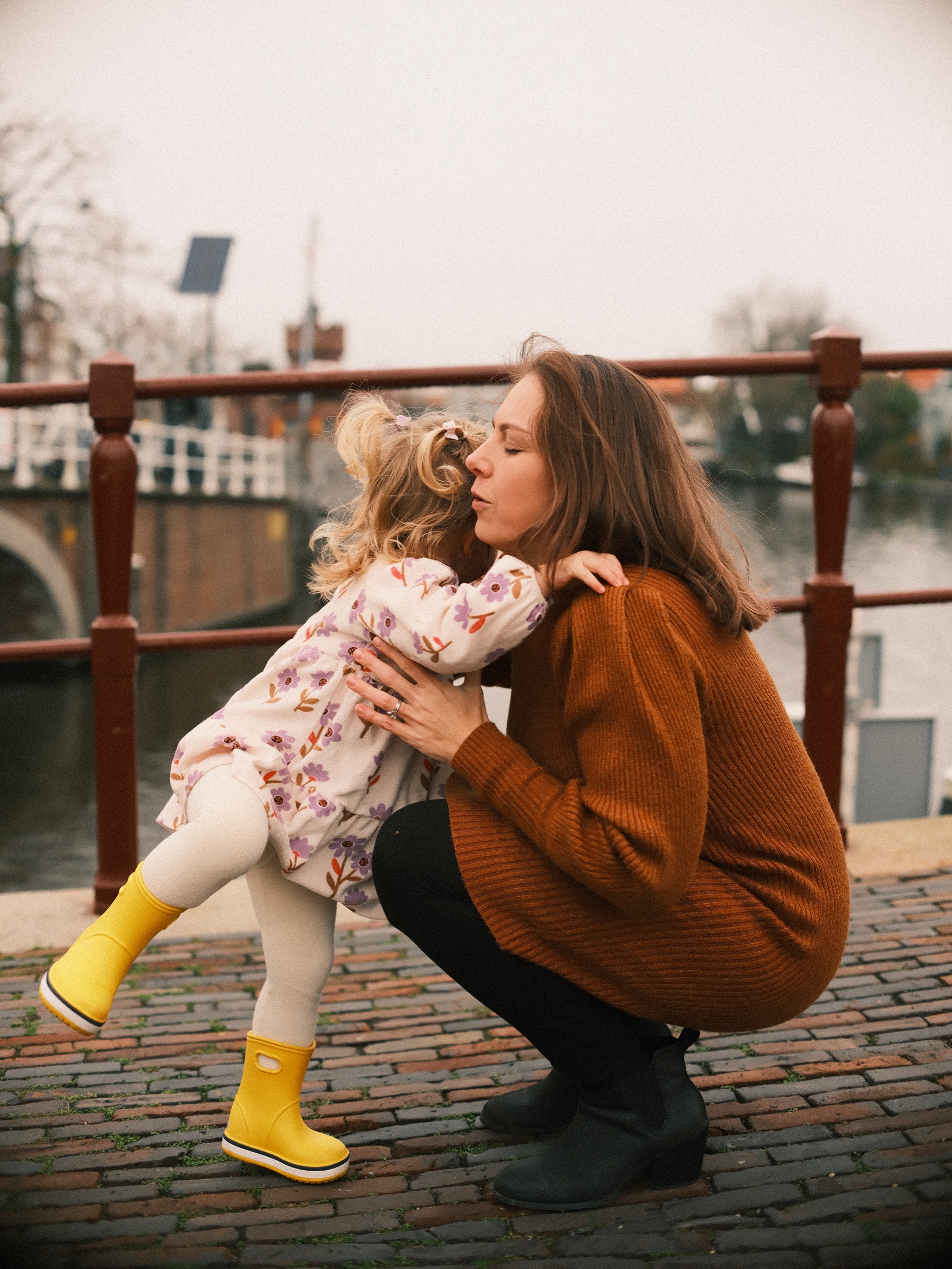 Peuter slaat haar armpjes om haar gehurkte moeder voor een kus bij de reling, met één gele laars omhoog