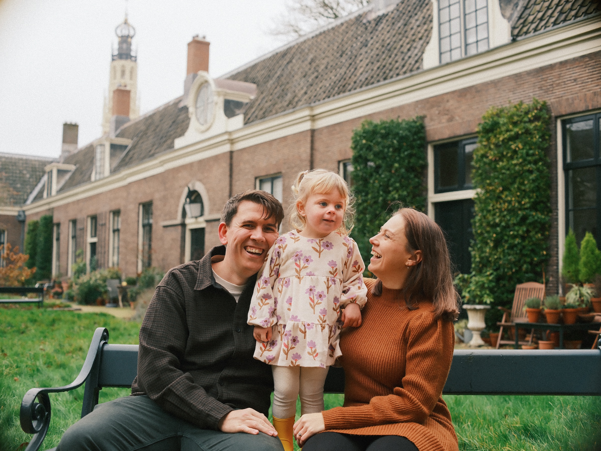 Op een bankje in een stil hofje staat de peuter tussen haar lachende ouders, met bakstenen huisjes en klimplanten erachter