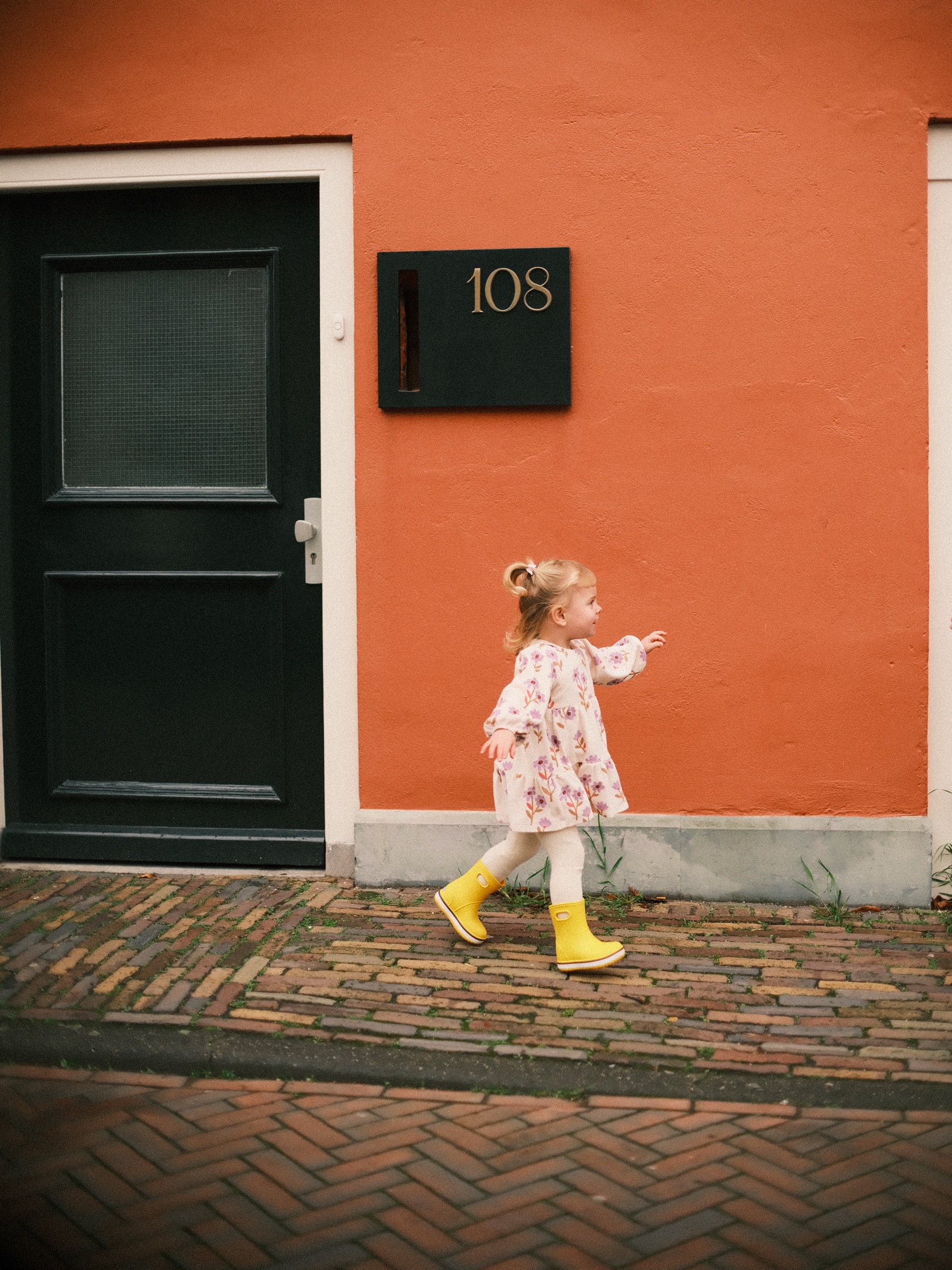 Meisje in een bloemenjurk en gele regenlaarzen loopt langs een zwarte deur in een oranje muur, met zwaaiende armpjes
