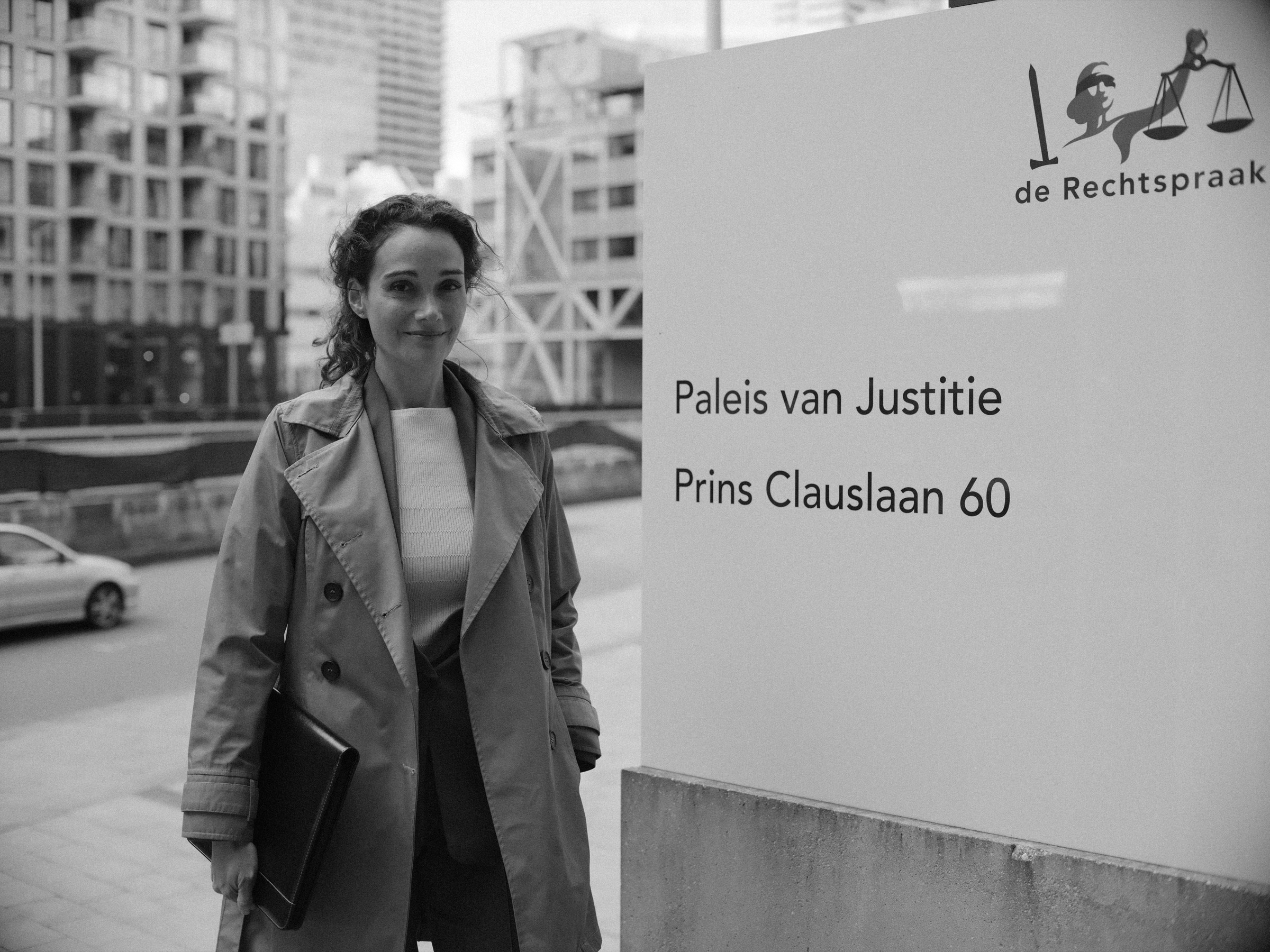 Vrouw in een trenchcoat lacht naast het bord van de Rechtspraak met Paleis van Justitie, Prins Clauslaan 60, een map onder haar arm