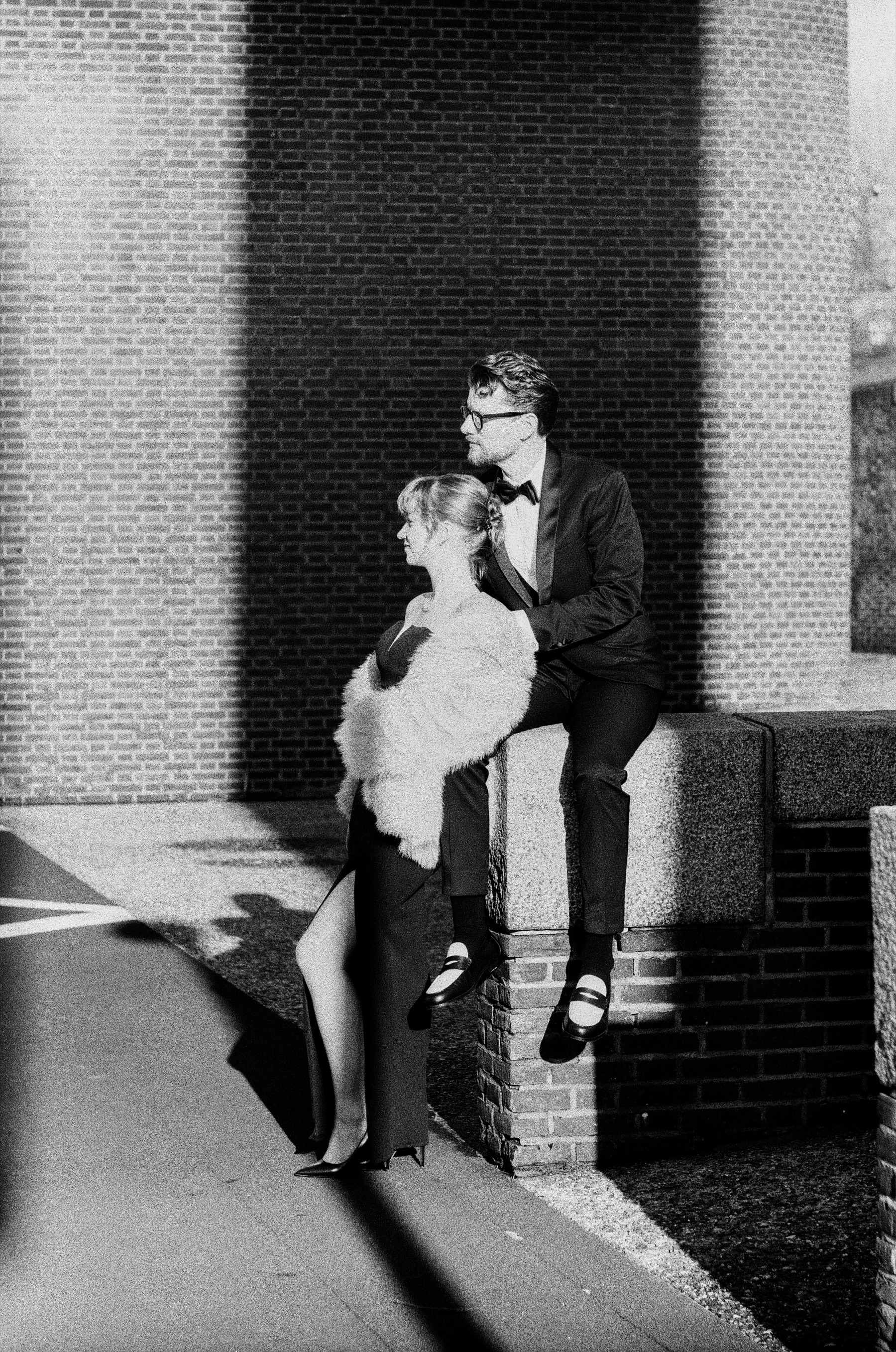 Man in a tuxedo sitting on a brick ledge as she leans back against him in a fur coat and a long slit dress, hard shadows on the wall