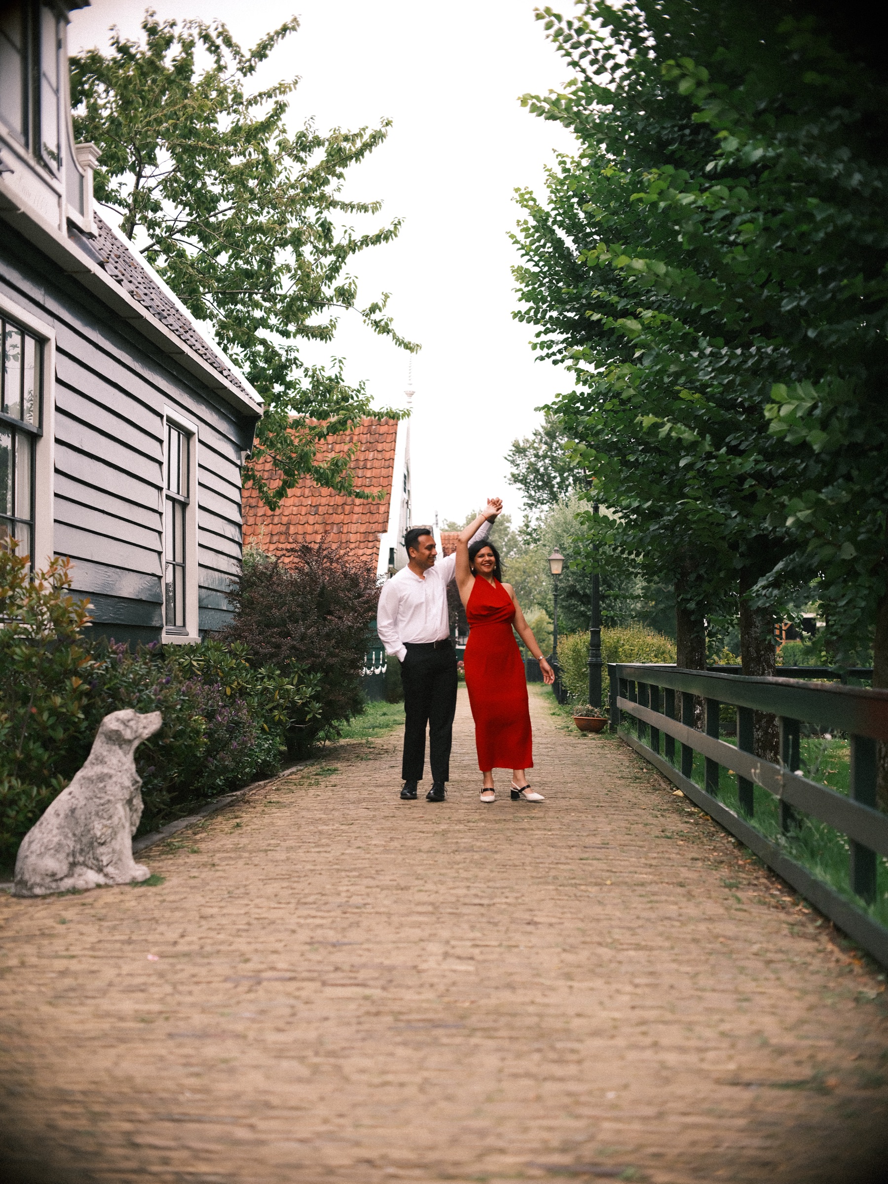 Man twirling his partner under his raised arm in a brick lane at the Zaanse Schans, a stone dog statue watching from the side