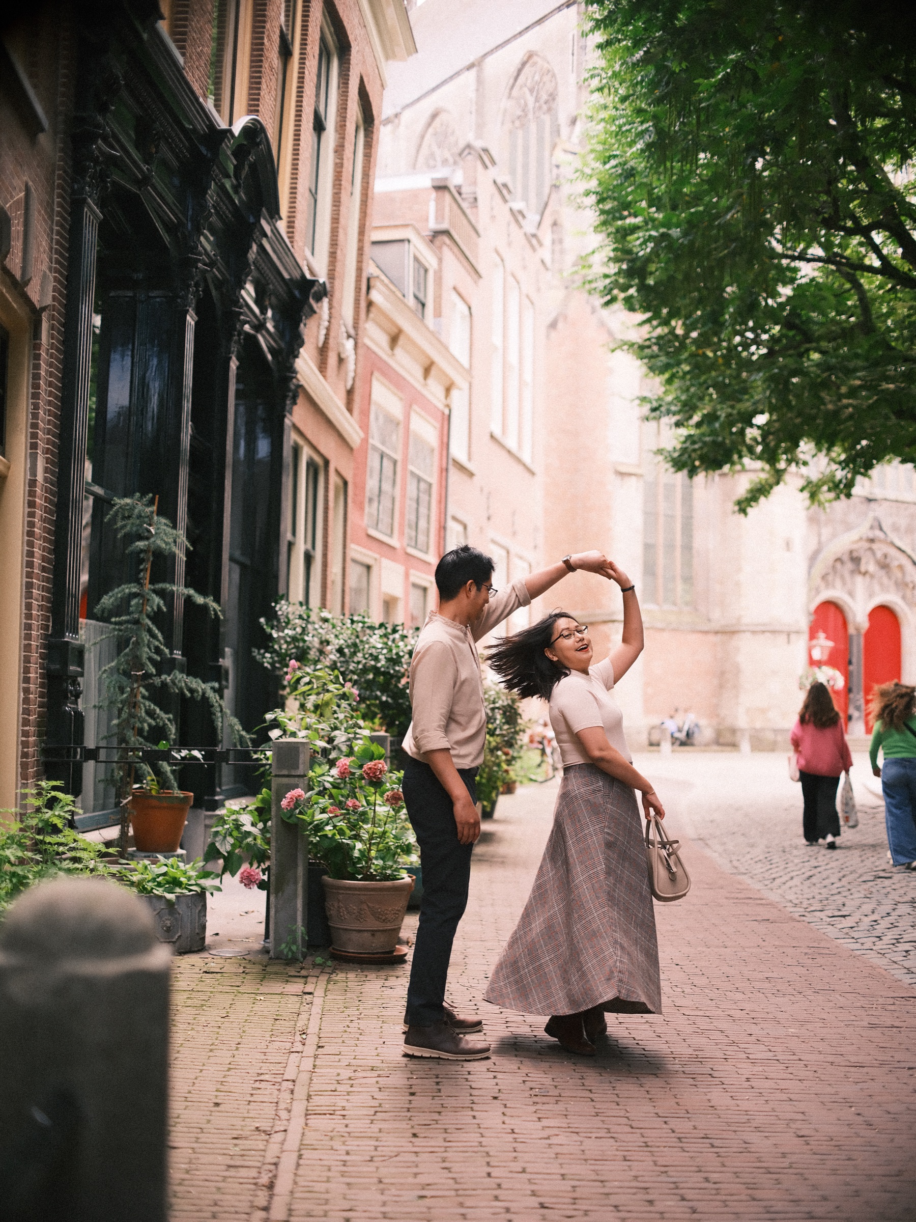 Mid-turn in the street, her dark hair flies as he lifts her hand, potted plants along the houses and the pale church behind