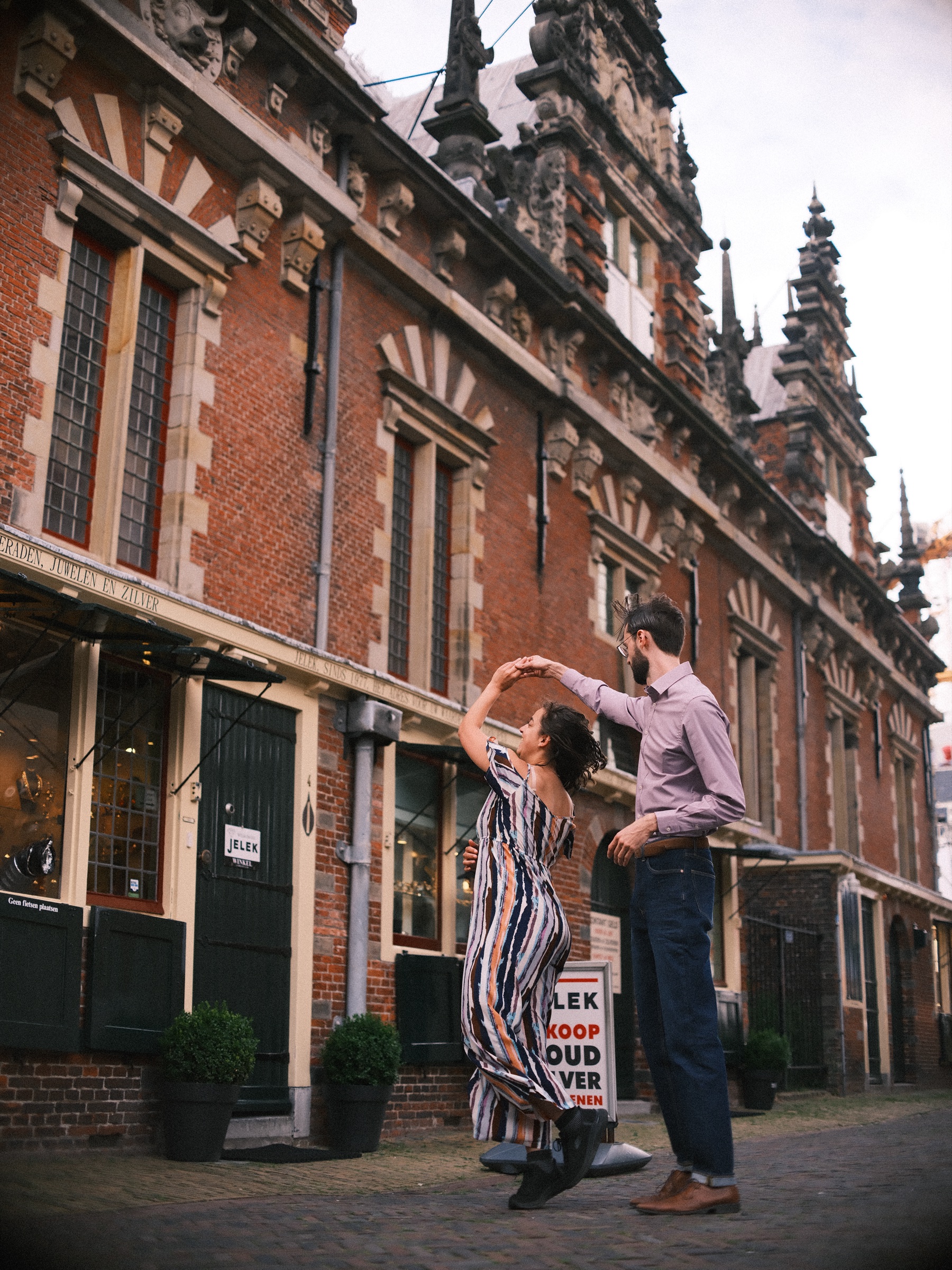 Man laat zijn vriendin in een gestreepte jumpsuit ronddraaien voor de Vleeshal, met hoge versierde gevels tegen een witte lucht