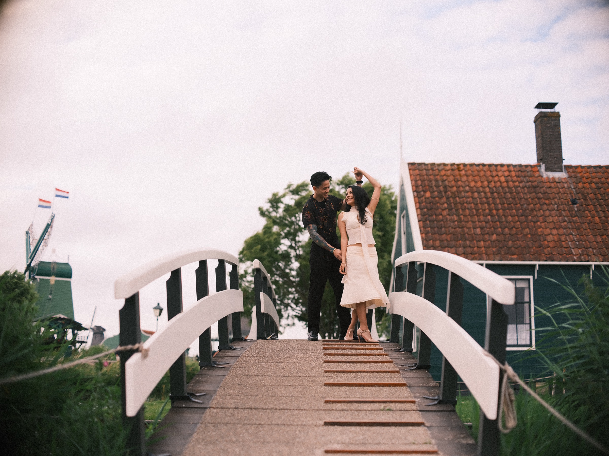 Couple dancing on a white wooden bridge, his raised hand twirling her, a green house with a red roof beside them at the Zaanse Schans