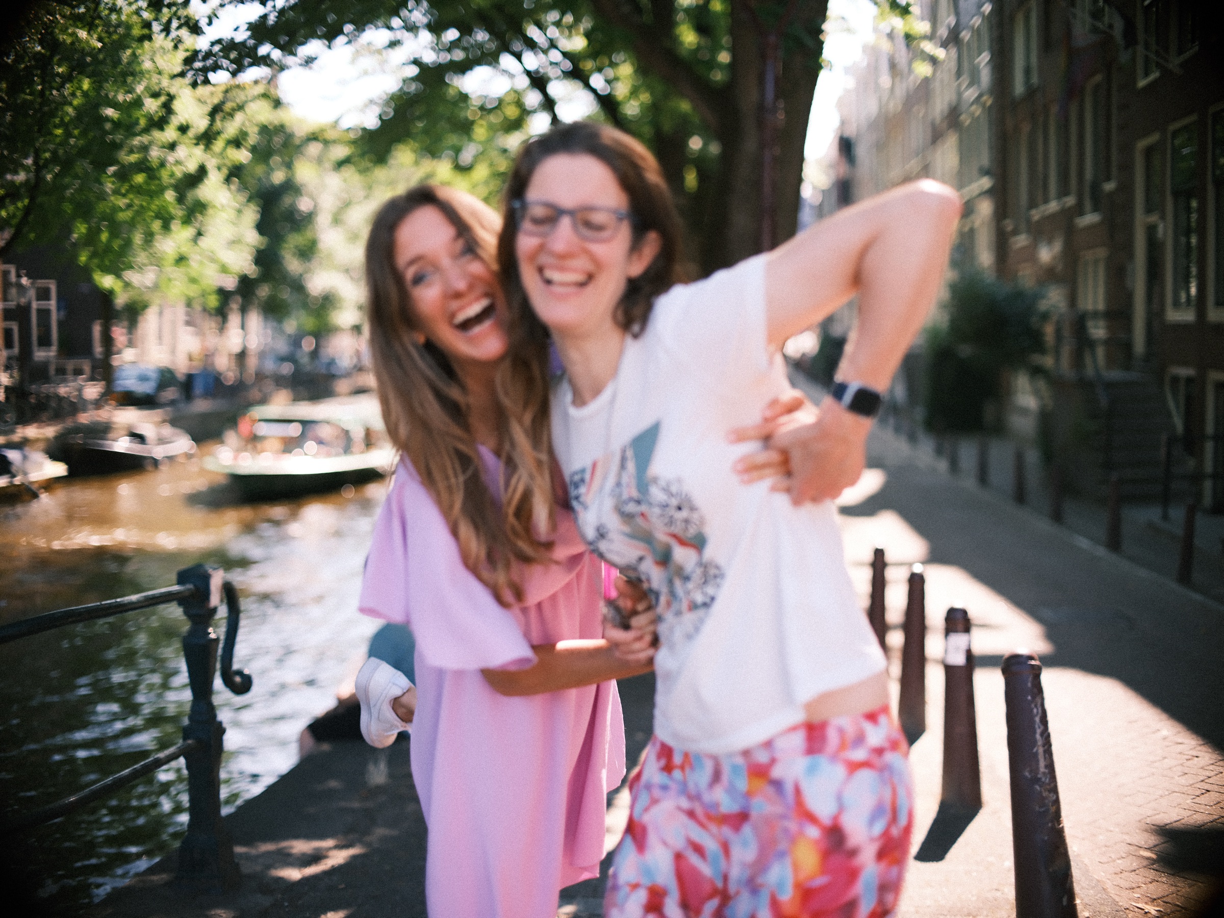 Two women laughing by a canal railing, one flinging out an arm, boats on the water and tall trees overhead