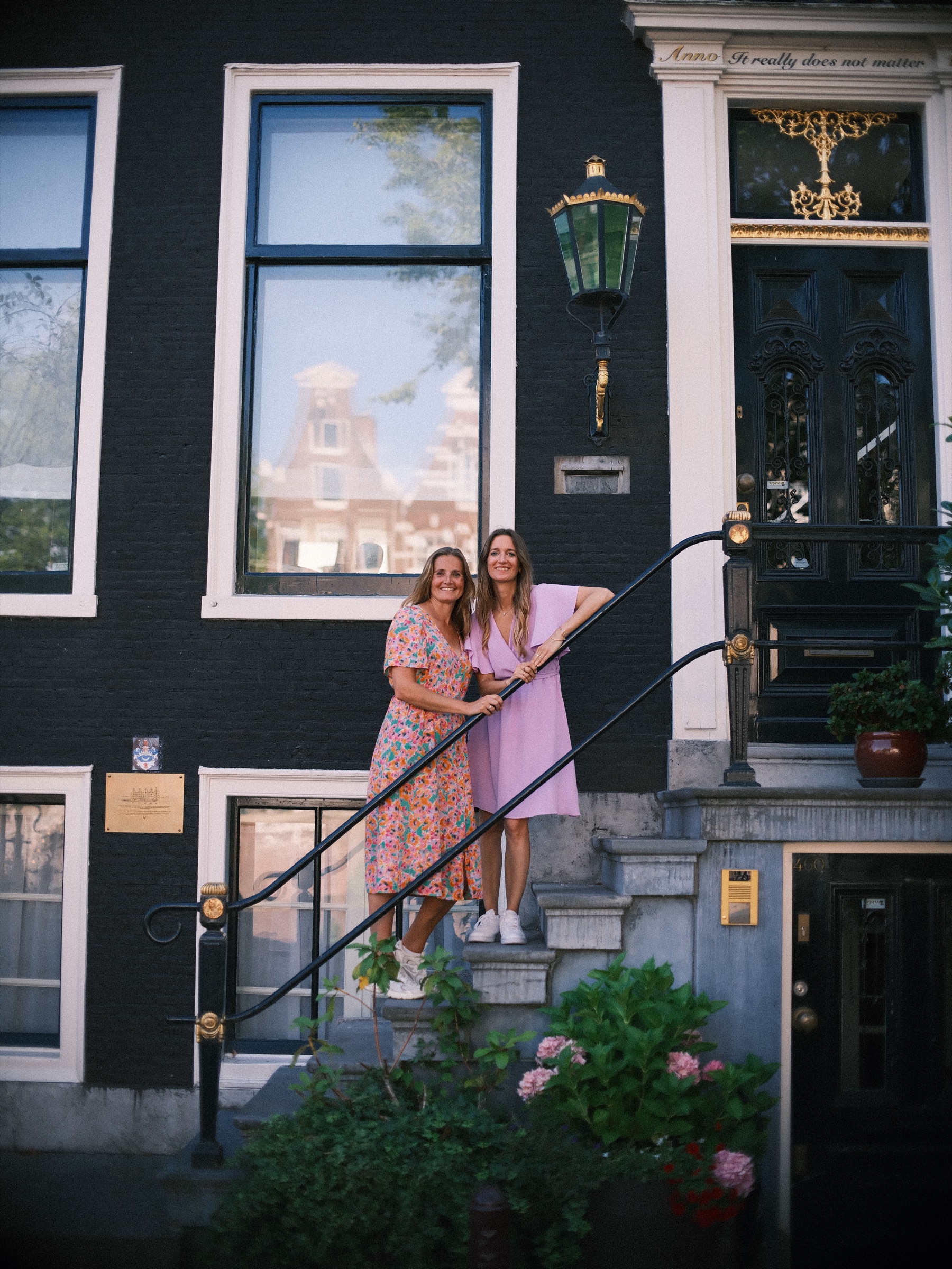 Two women standing on the stoop of a dark canal house with white window frames, holding the black iron railing