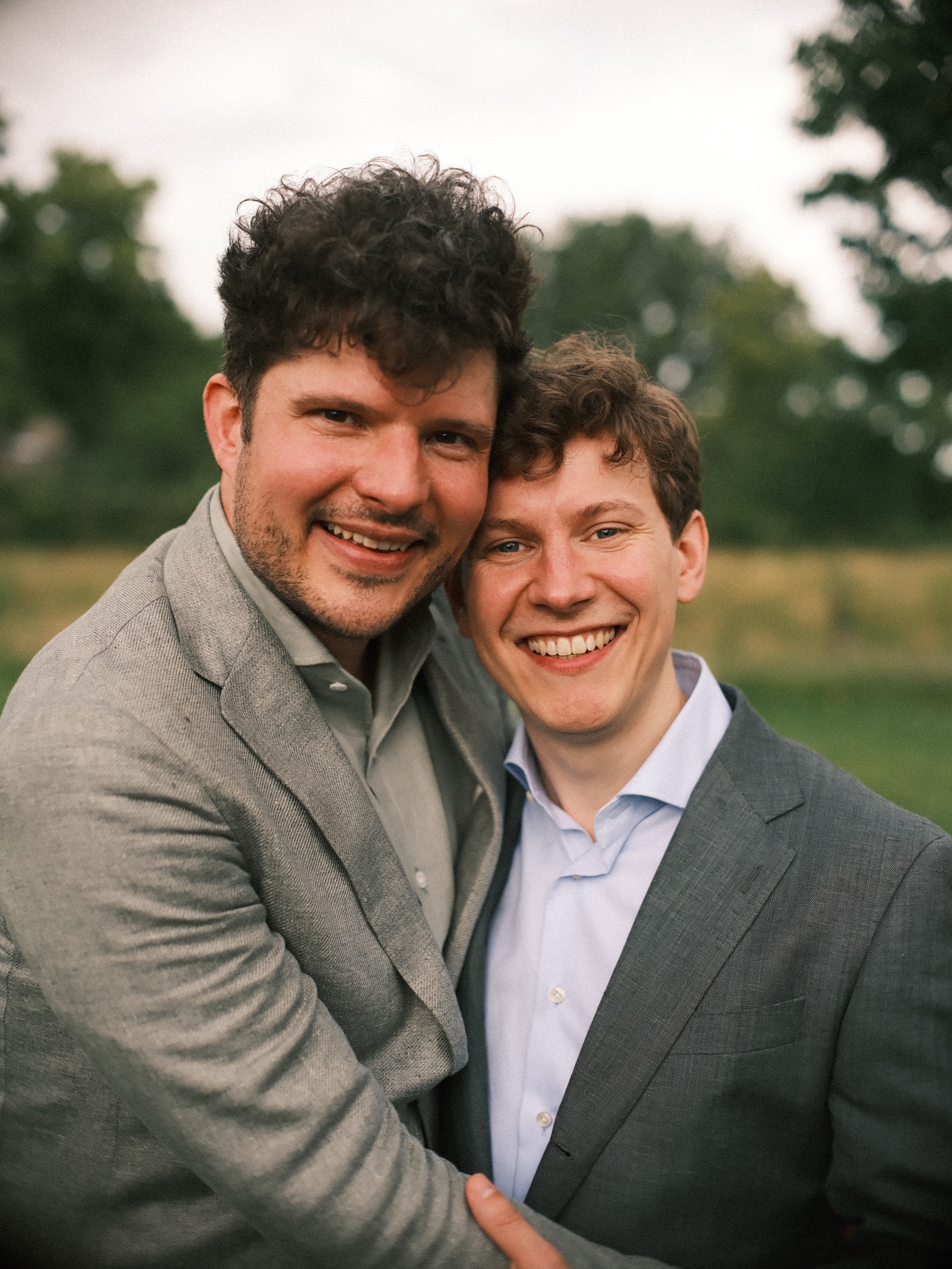 Cheek to cheek and beaming, two grooms in a linen jacket and a gray suit pose on the grass just after their same-sex wedding