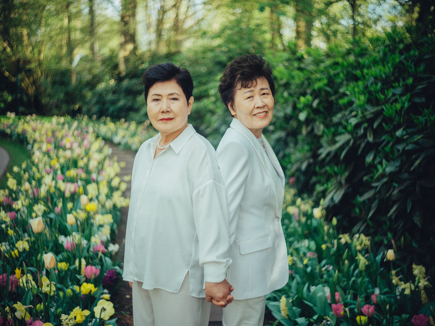 Twee dames van de oudste generatie, allebei in het wit, houden elkaars hand vast tussen zachtgele tulpen in Keukenhof