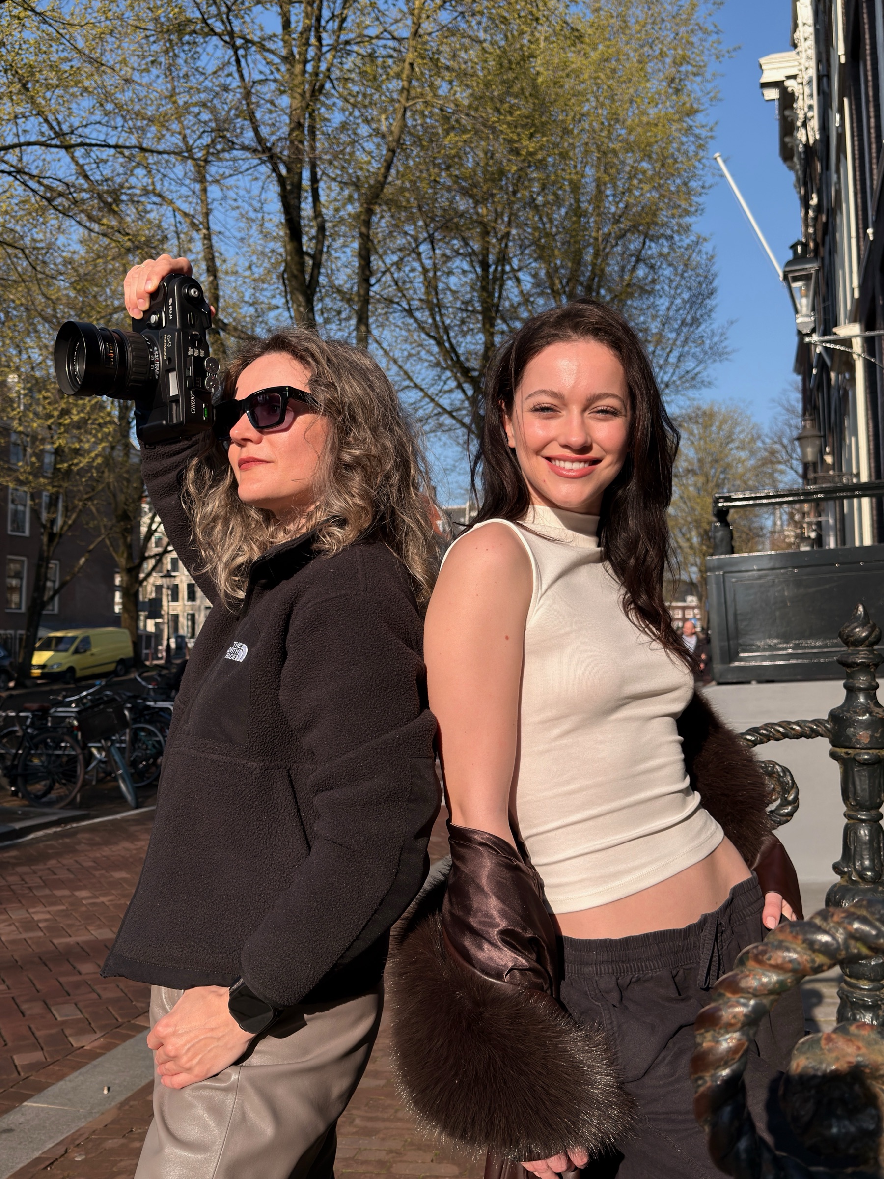 Lachende vrouw in een wit topje en een bruine jas naast een vrouw met zonnebril die een analoge camera omhoog houdt op een zonnige brug