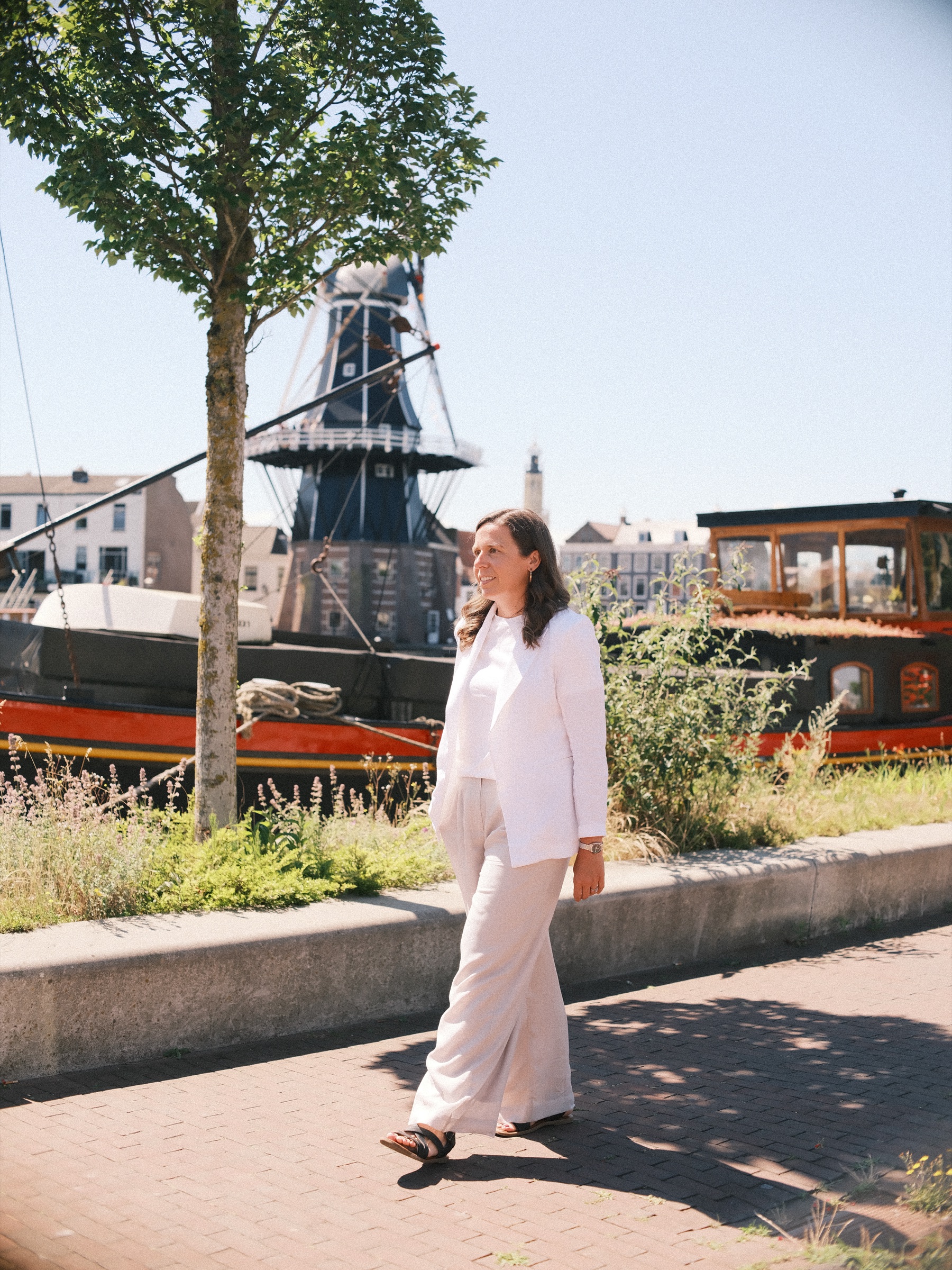 Vrouw in het wit loopt over het pad langs een oud schip, met molen De Adriaan achter een jonge boom aan het Spaarne