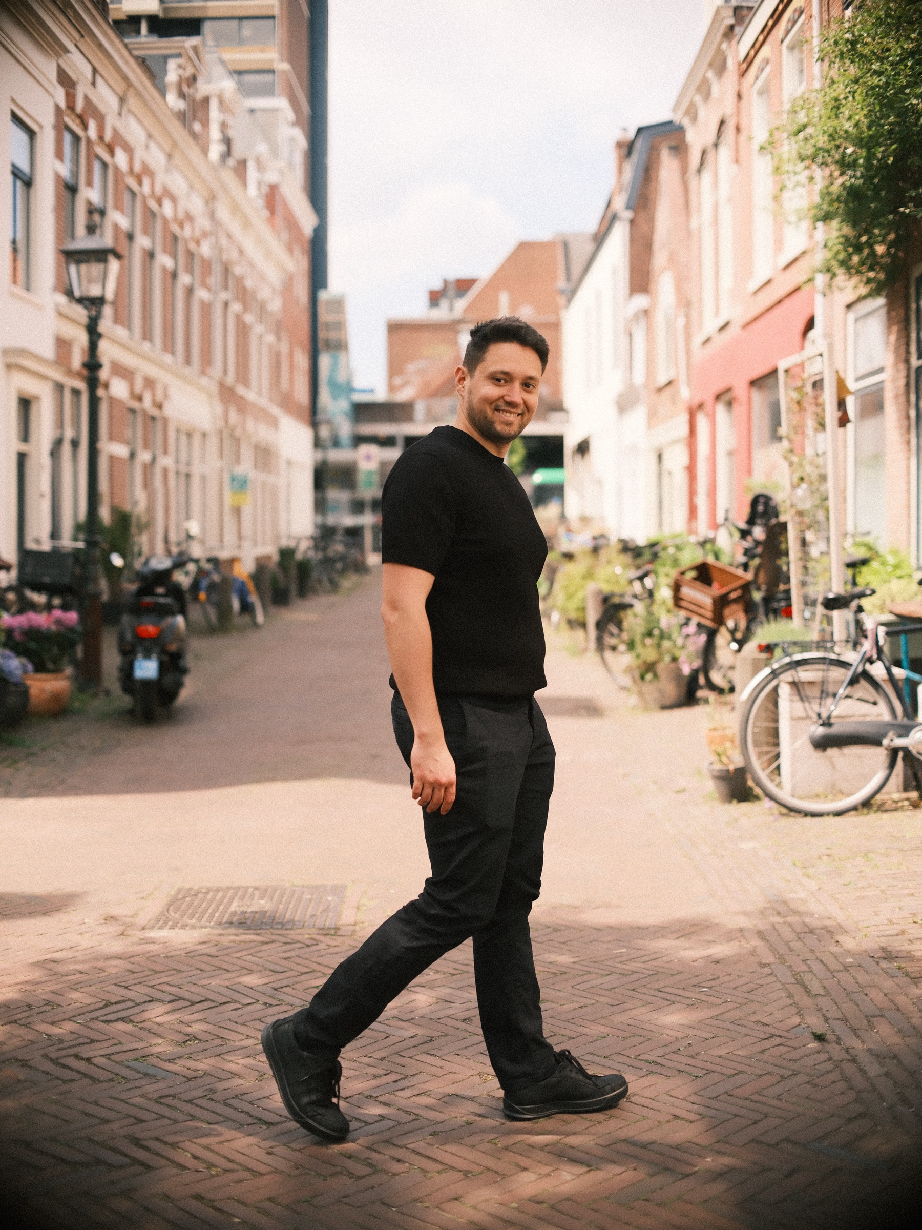 Man in zwart T-shirt steekt een klinkerstraat in Haarlem over, met fietsen, planten en smalle huizen in de zon achter hem