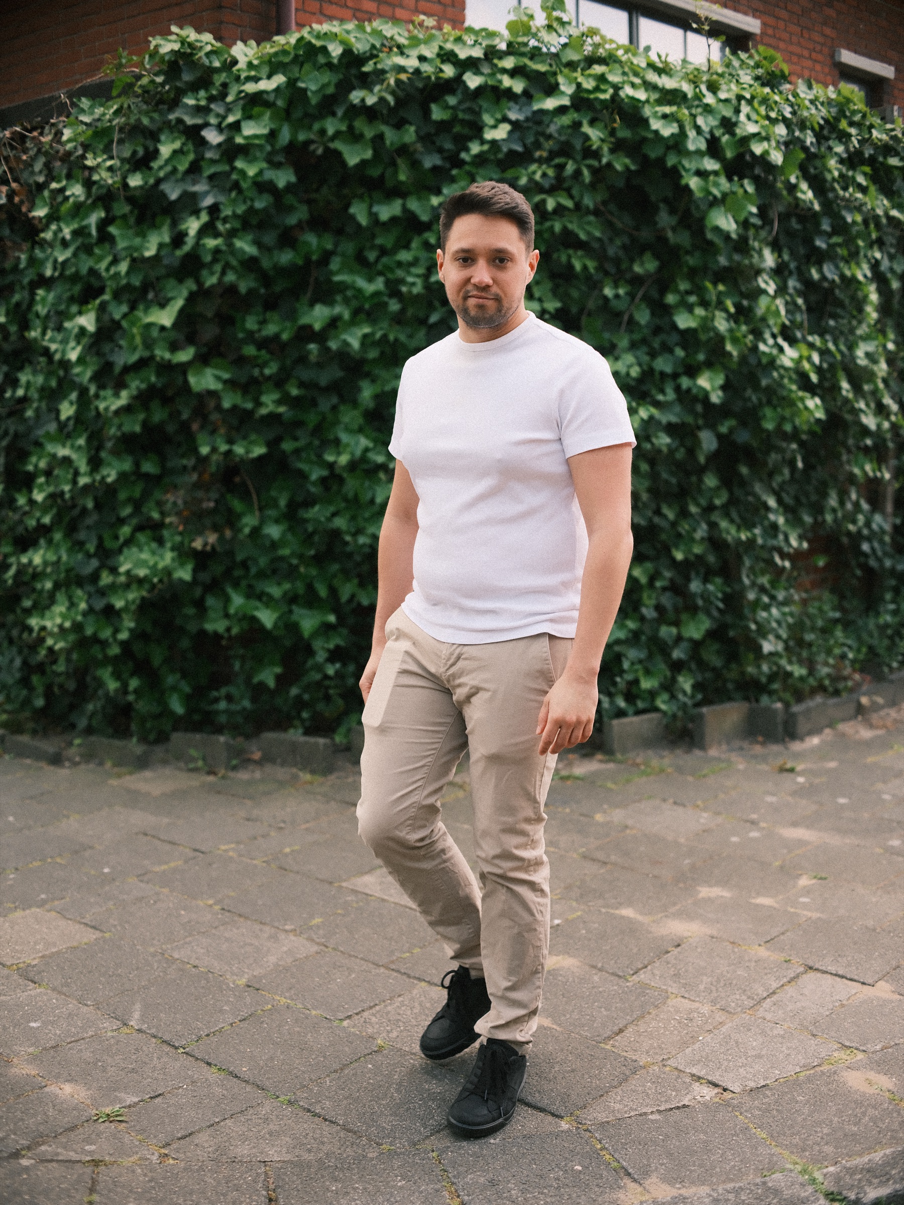 Man in wit T-shirt en beige broek loopt naar de camera toe, voor een muur vol klimop en een gebouw van rode baksteen