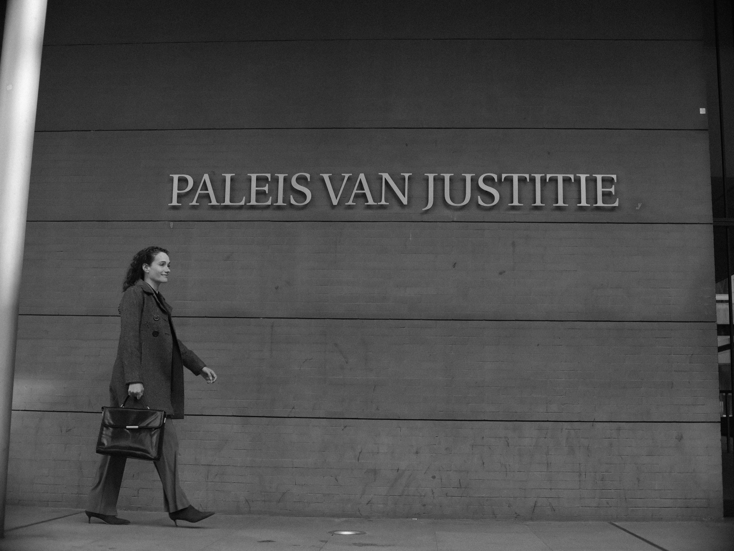 Vrouw in een lange donkere jas loopt met een leren tas langs de gevel van het Paleis van Justitie, met de naam in grote letters erboven