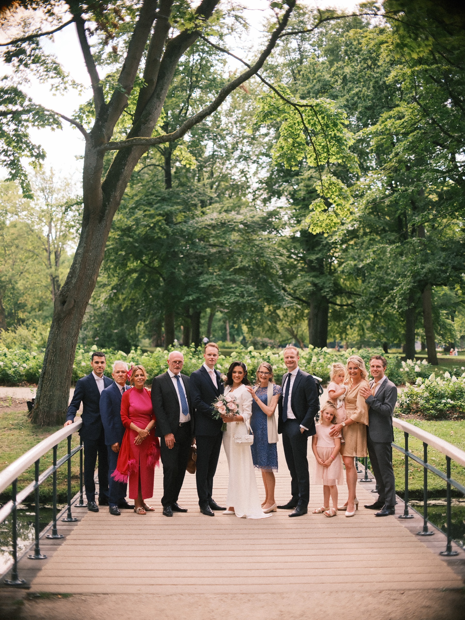 Grote groep van twee families op een wit bruggetje in het park, met hoge bomen en witte bloemen erachter