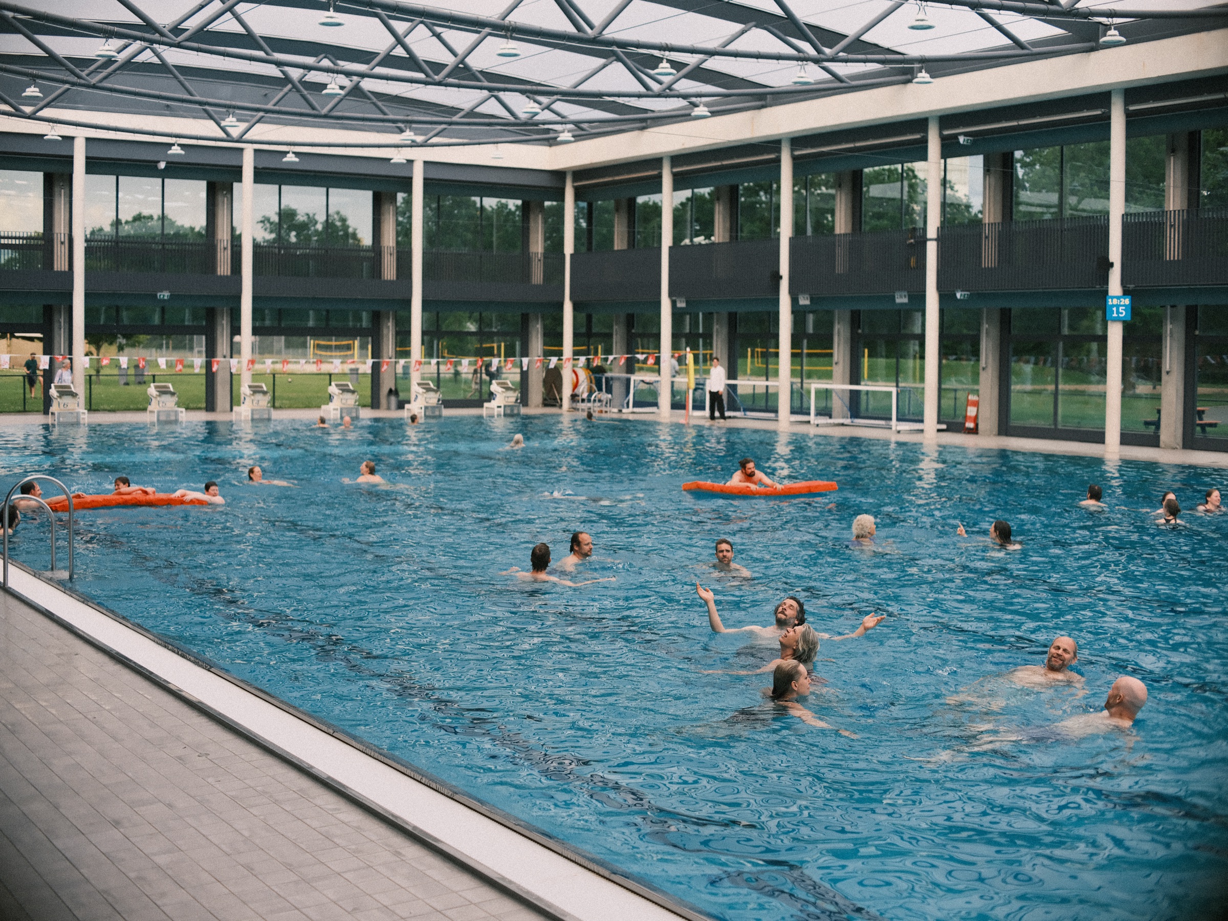 Wedding guests swimming in a long pool under an open steel roof frame in Utrecht, orange floats drifting on the water