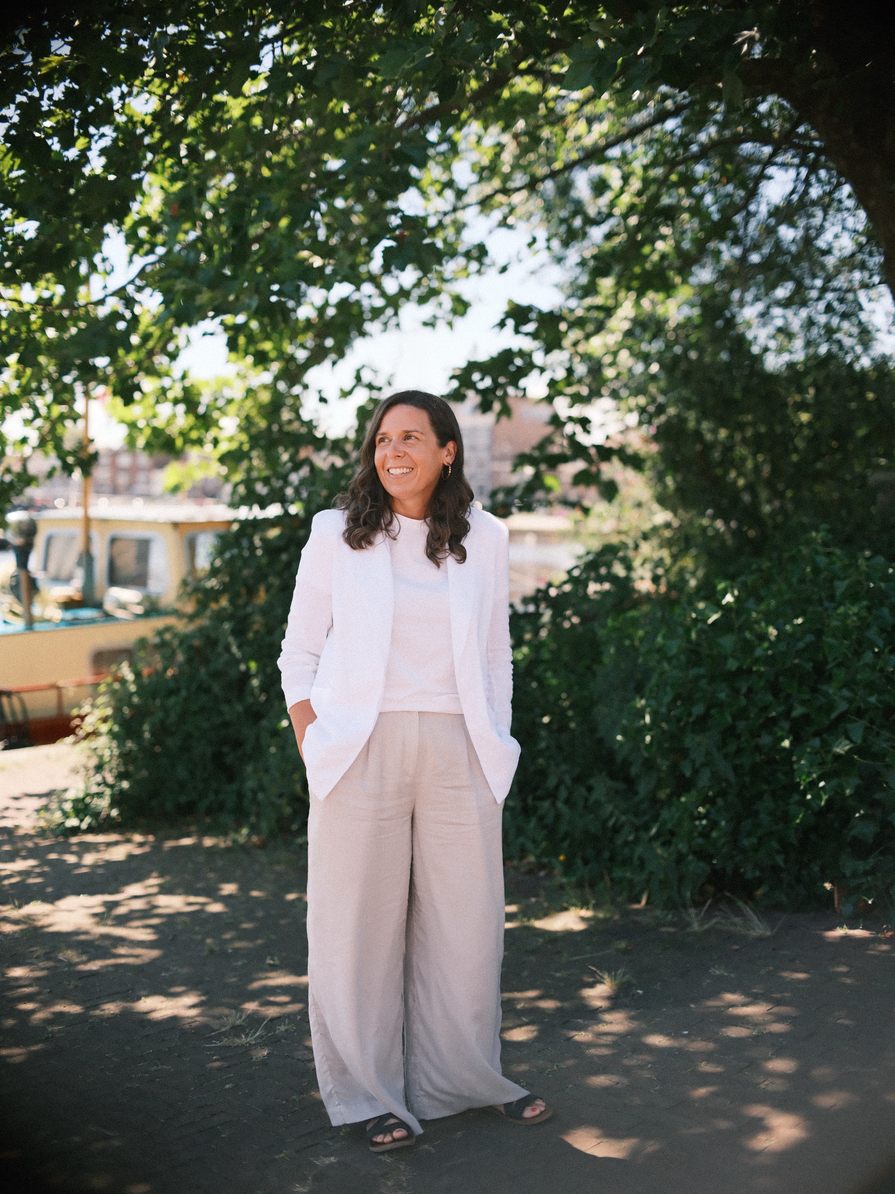 Vrouw in witte blazer onder een bladerrijke boom, handen in haar zakken, met de rivier en huizen tussen het groen door