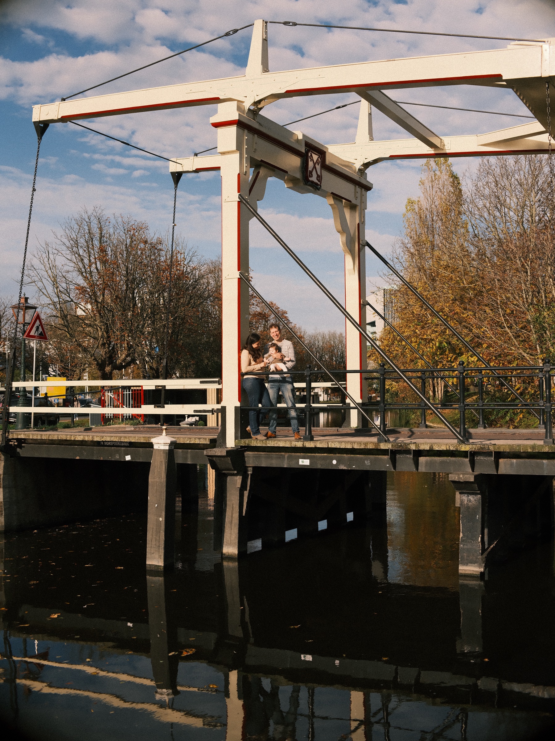 White wooden drawbridge over dark water on a sunny autumn afternoon in Leiden, the family small at its railing between golden trees