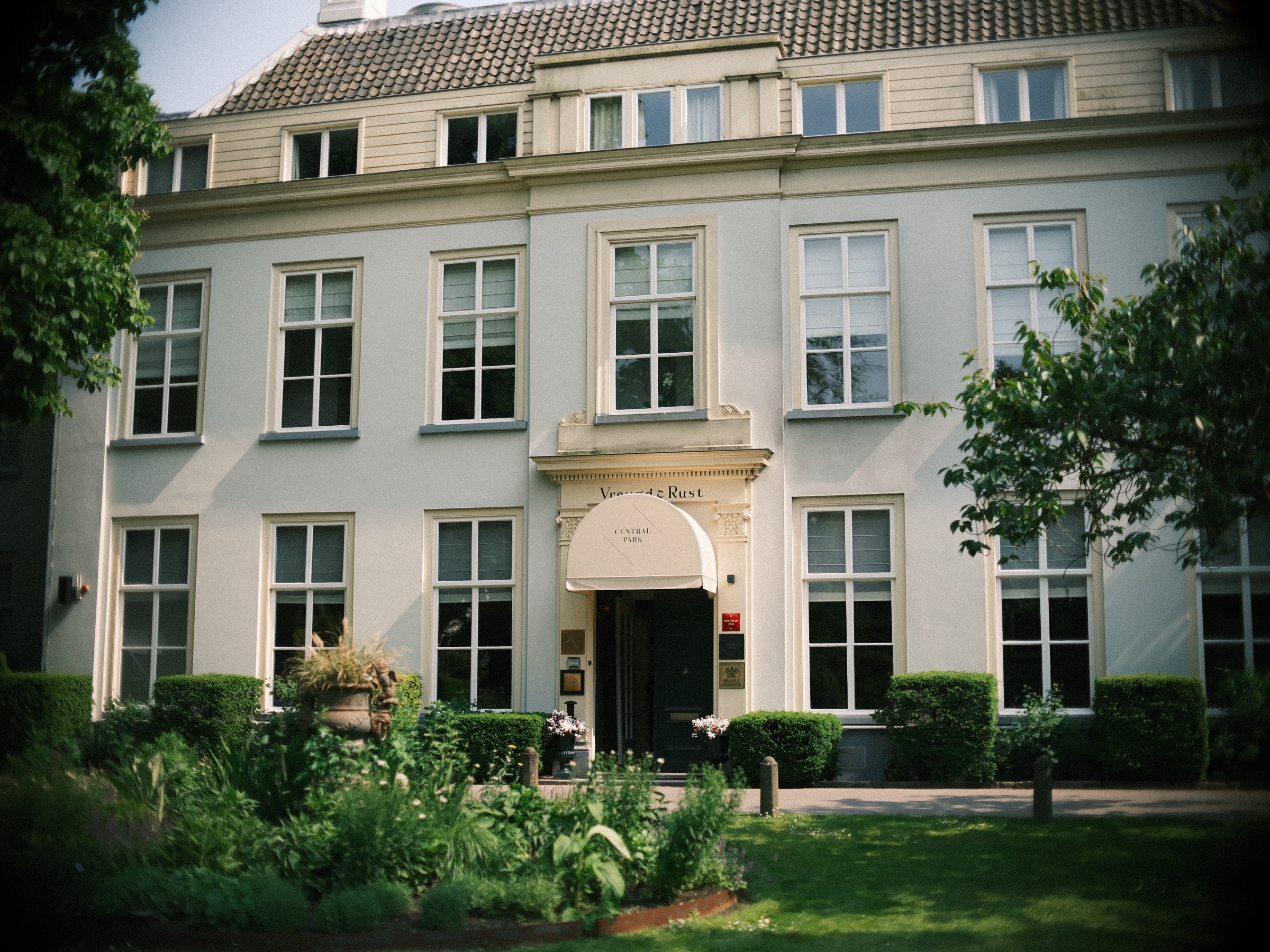 Witte villa op landgoed Vreugd en Rust in Voorburg met een crèmekleurige luifel boven de deur, een gazon en bomen in de zon