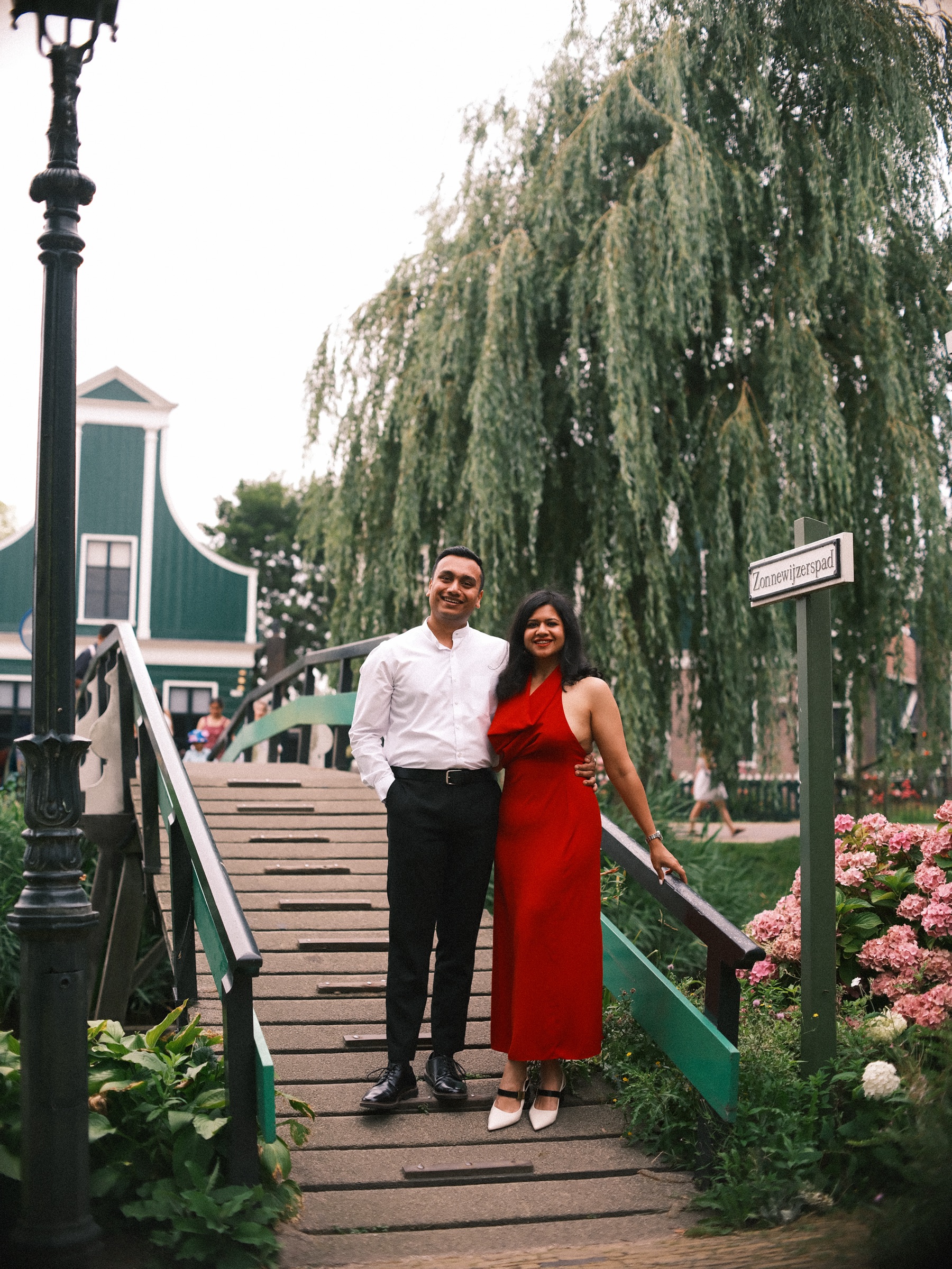 Couple on a wooden footbridge with green railings under a weeping willow, a lamppost, pink hydrangeas and the Zonnewijzerspad sign beside them